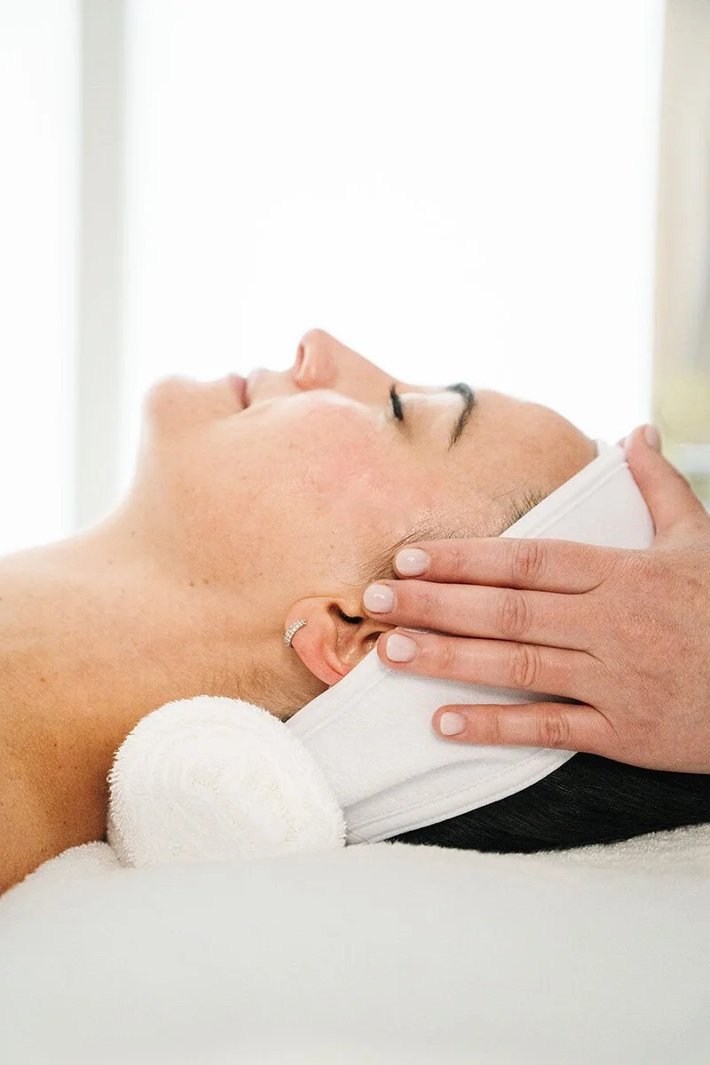 A woman receiving a facial massage or treatment at a spa, lying down with her head supported by a rolled towel, eyes closed, and a white headband with a towel wrapped around her head.