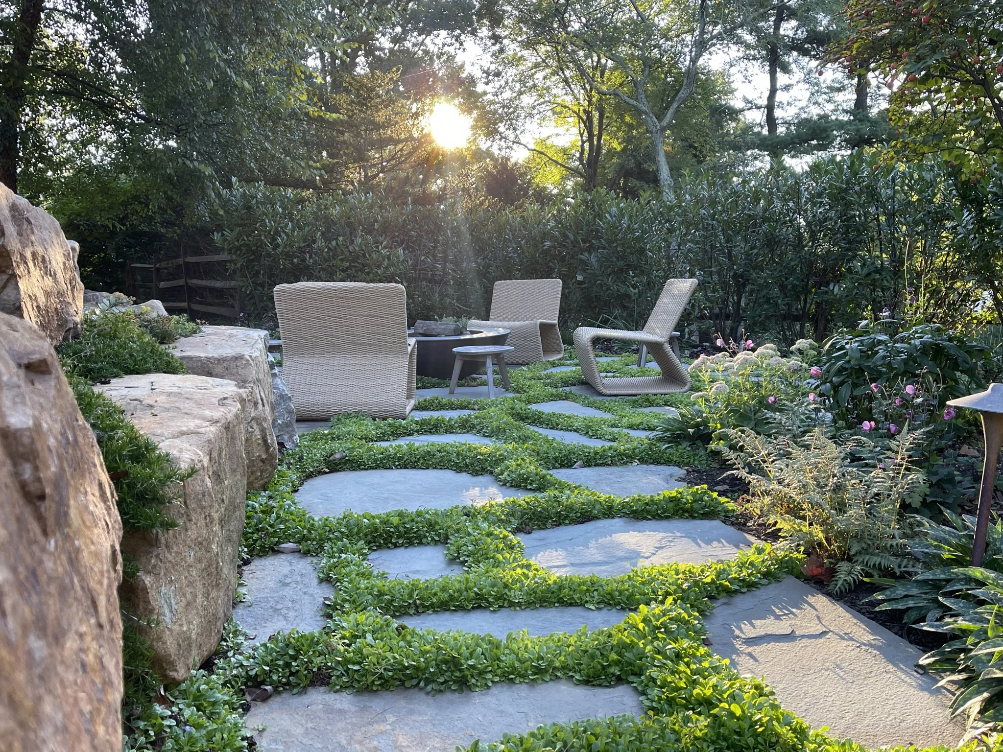 Garden patio with stone walkway, wicker chairs, and surrounding greenery.