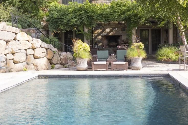 Backyard with a swimming pool, stone wall, and patio area featuring lounge chairs, potted plants, and greenery.