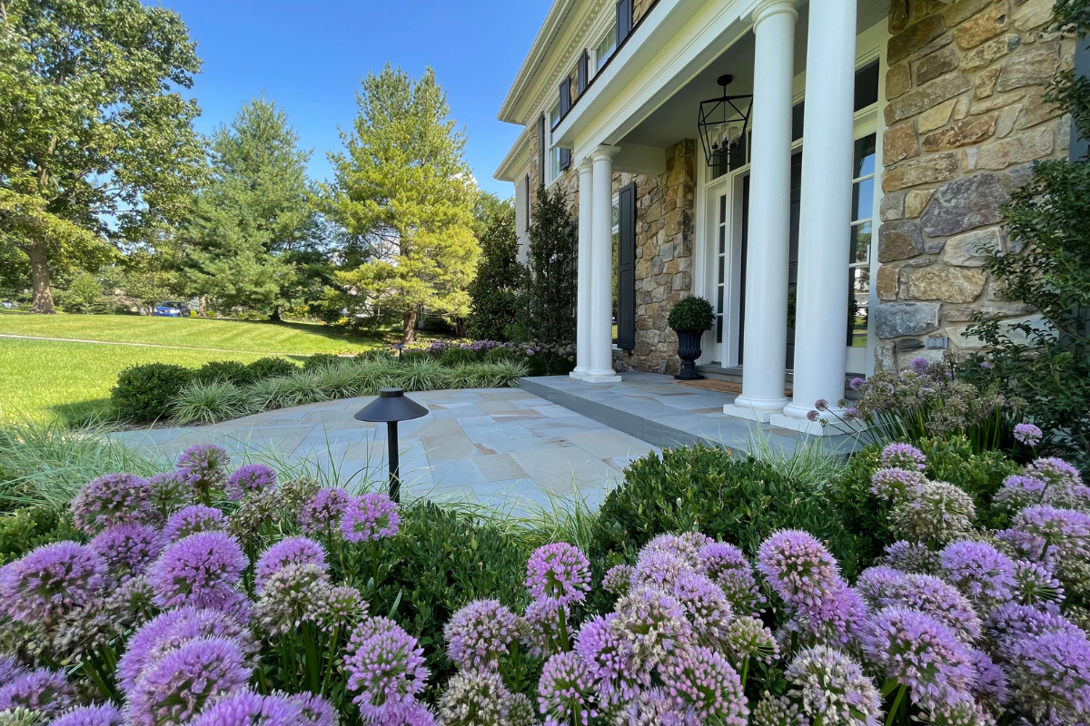 Front porch of a house with stone facade, white columns, and a manicured garden featuring purple flowers and greenery on a sunny day.