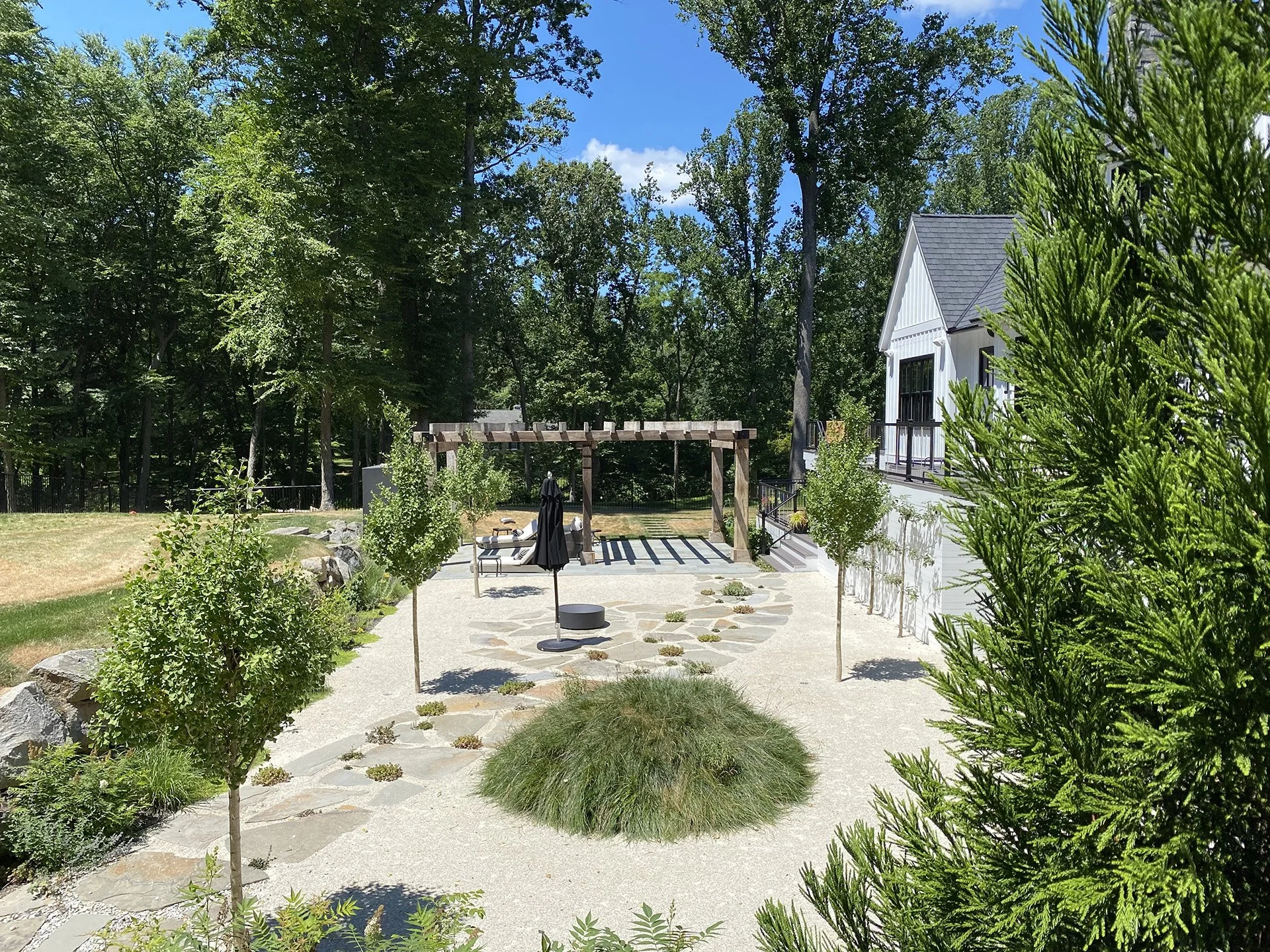 A landscaped garden with small trees, stone pathways, and seating area under a pergola, surrounded by lush green trees.