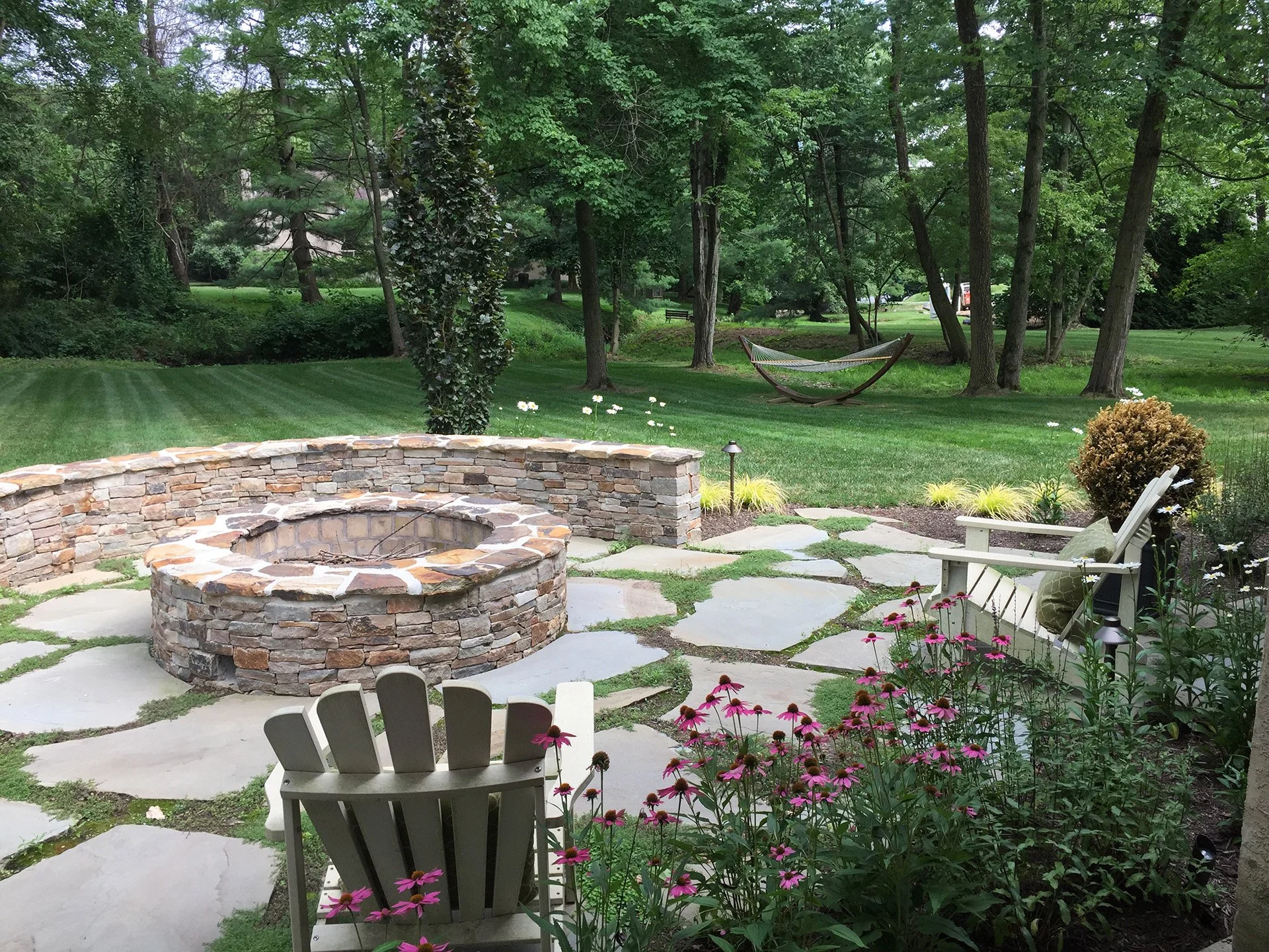 Backyard with stone fire pit, Adirondack chairs, hammock, and flowering plants. Lush green lawn and trees surround the patio.