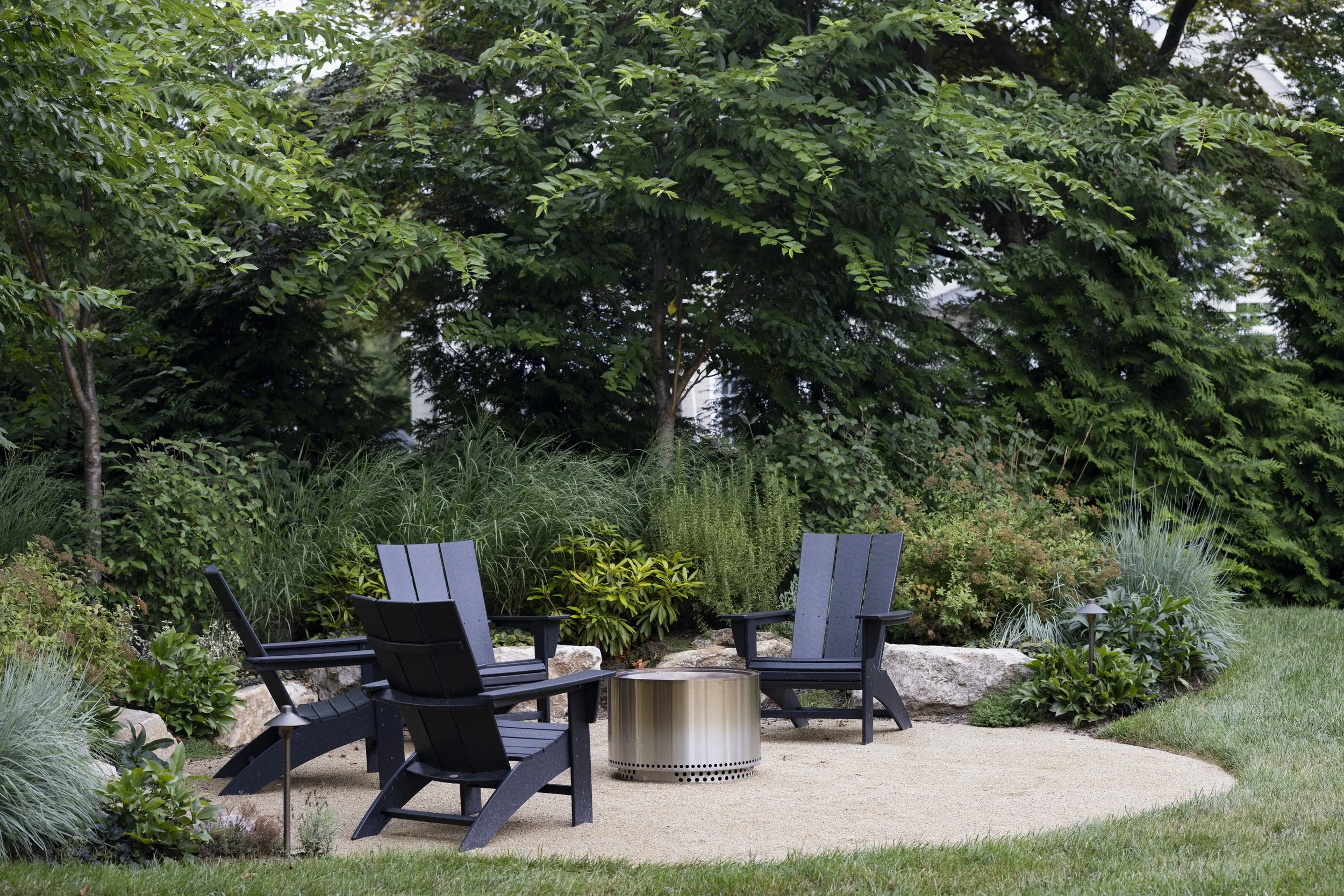 Outdoor seating area with black Adirondack chairs and a fire pit surrounded by lush greenery and trees.