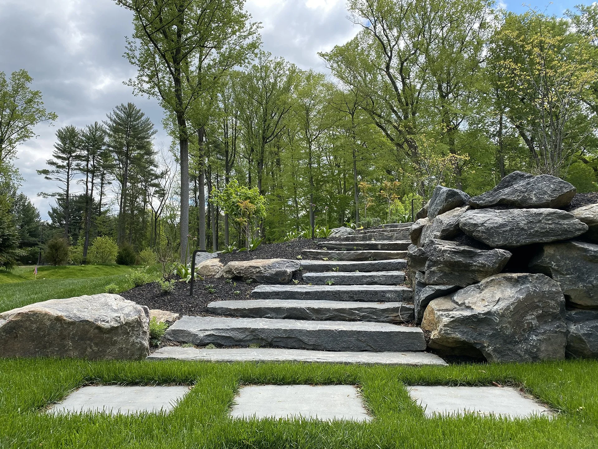 Stone steps leading up a grassy hill with large rocks on the side, surrounded by trees in a lush, green natural setting.