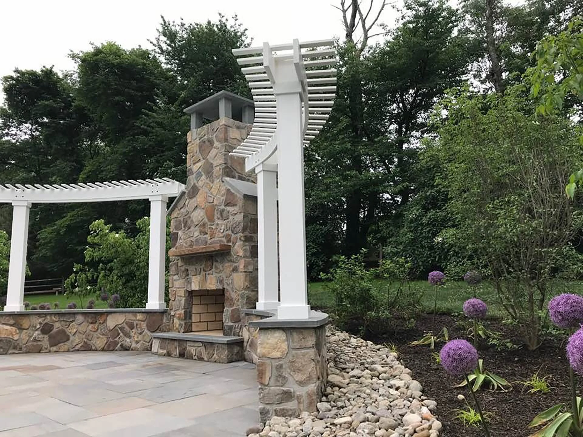 Outdoor stone fireplace with pergola and garden featuring purple flowers and trees.