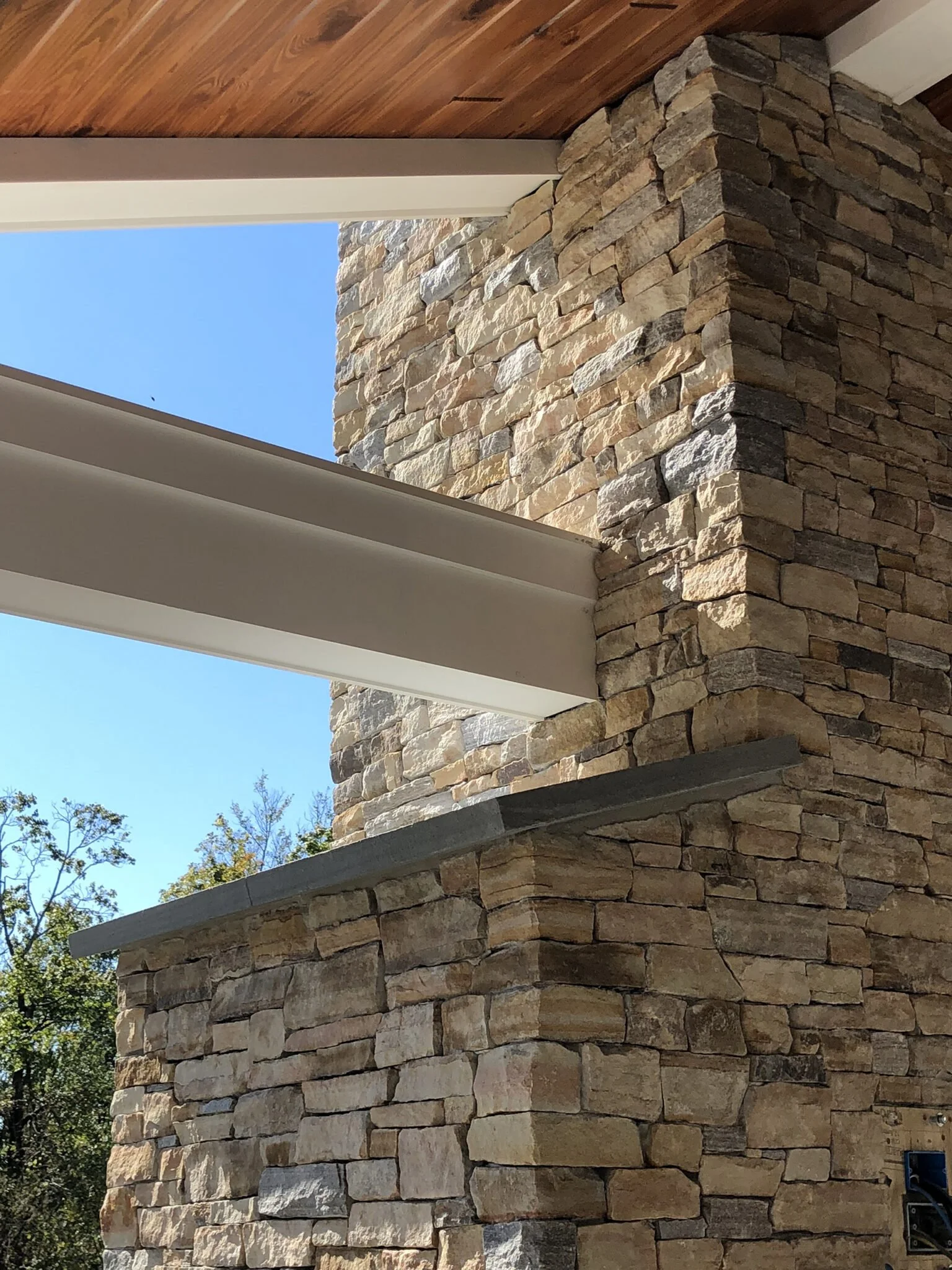 Rustic stone column supporting wooden roof beams and ceiling, surrounded by trees and a blue sky.