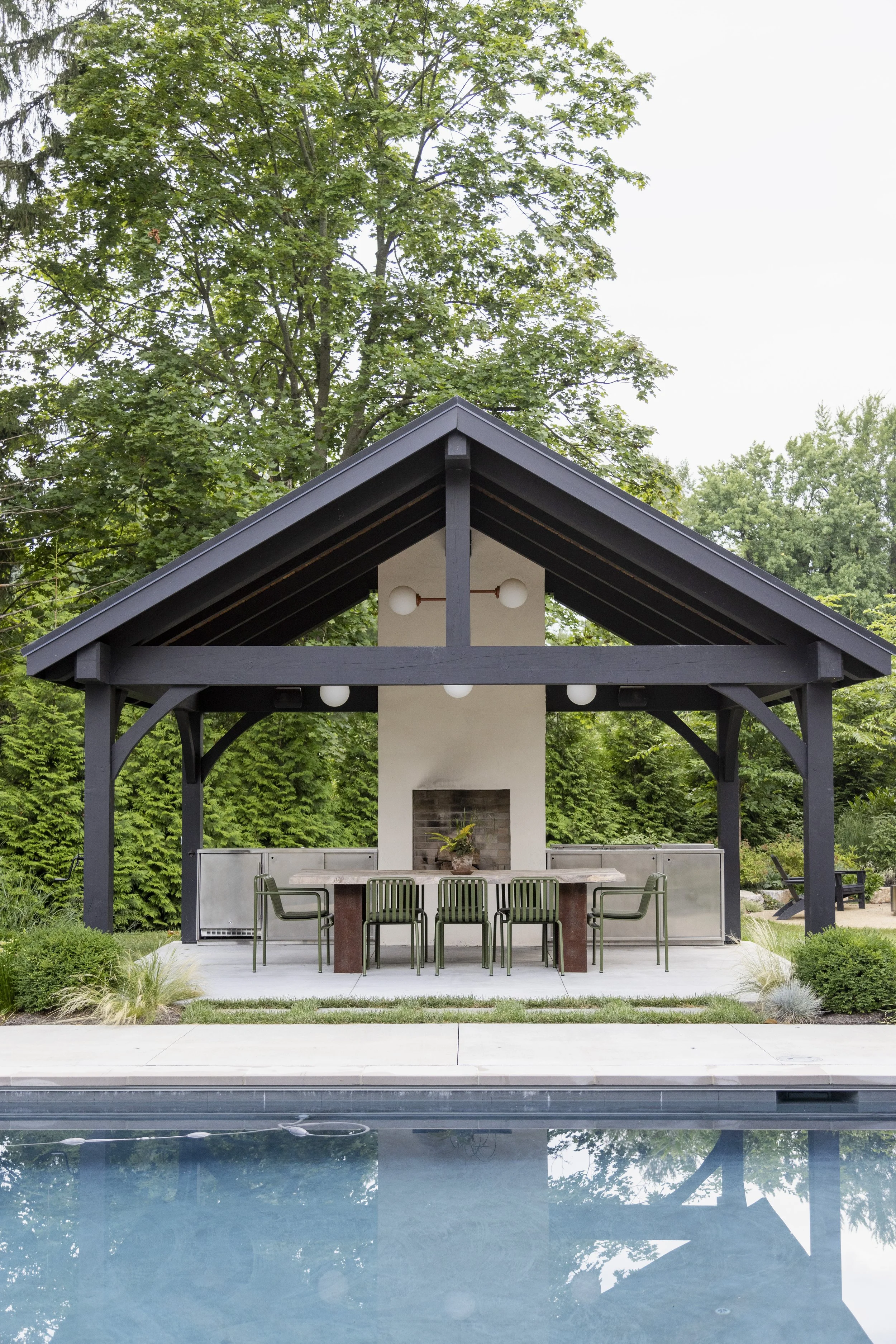 Outdoor poolside pavilion with a modern, open-air design, featuring a fireplace, dining table, green chairs, and overhead lighting, surrounded by lush green trees and landscaping.