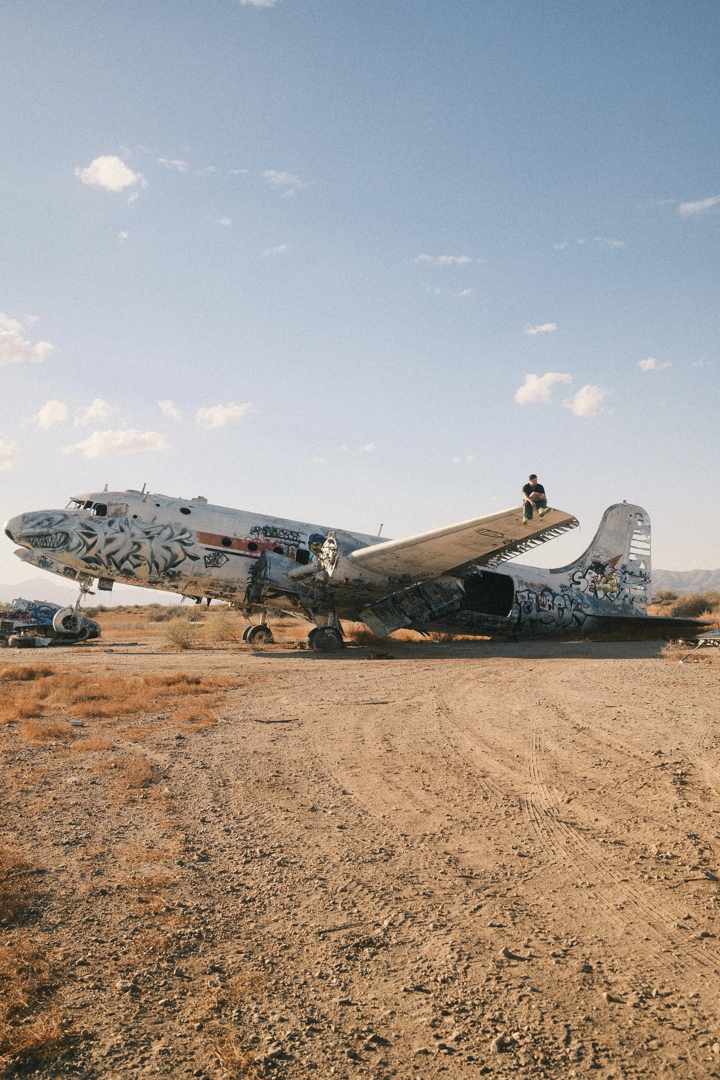 An abandoned airplane covered in graffiti in a desert landscape with a person sitting on top of the wing during daytime.