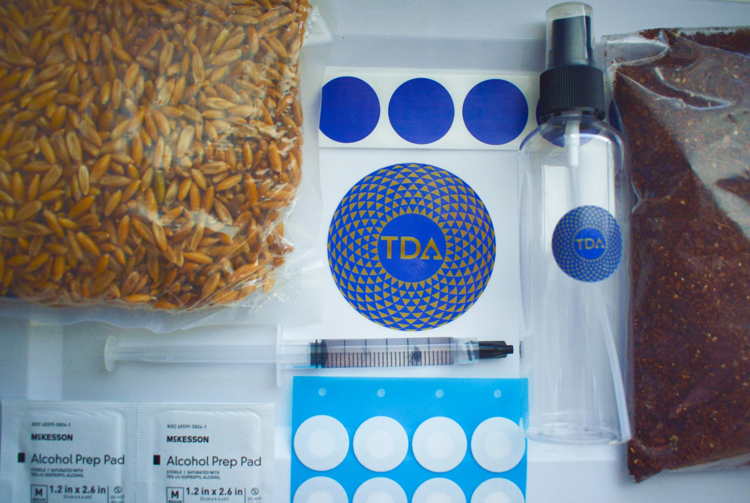 A collection of materials for testing or analysis, including plastic bags of grains or seeds, alcohol prep pads, a syringe, a spray bottle, circular stickers with the label 'TDA', and other sealed samples.