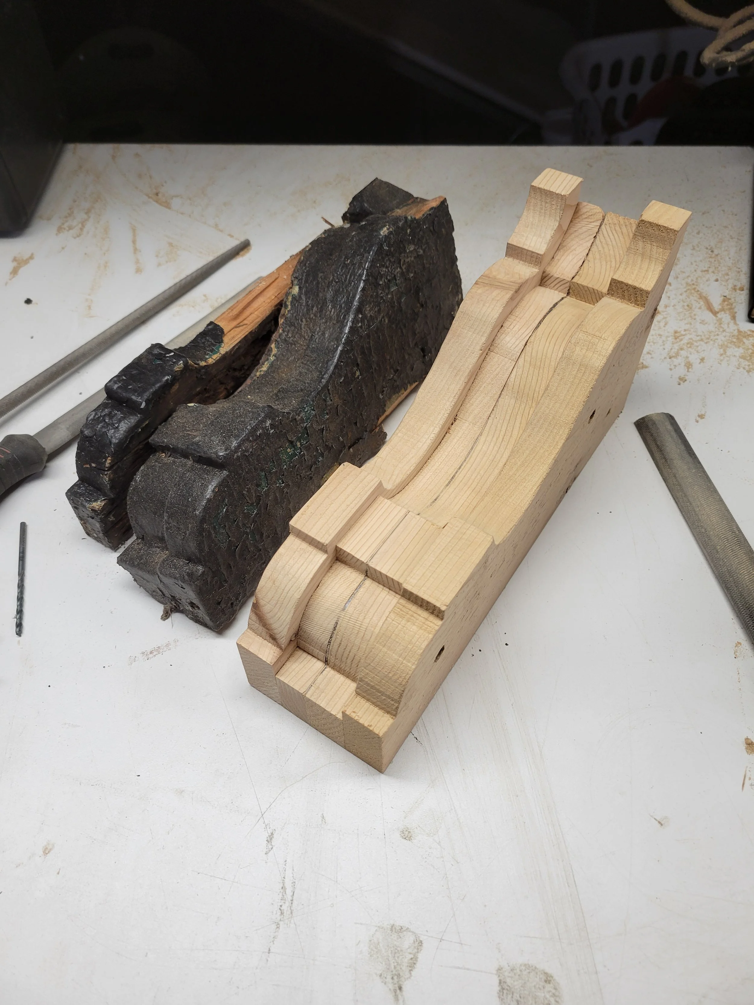 A woodworking workspace with a piece of carved light-colored wood, a dark burnt block of wood, and various woodworking tools on a white table.