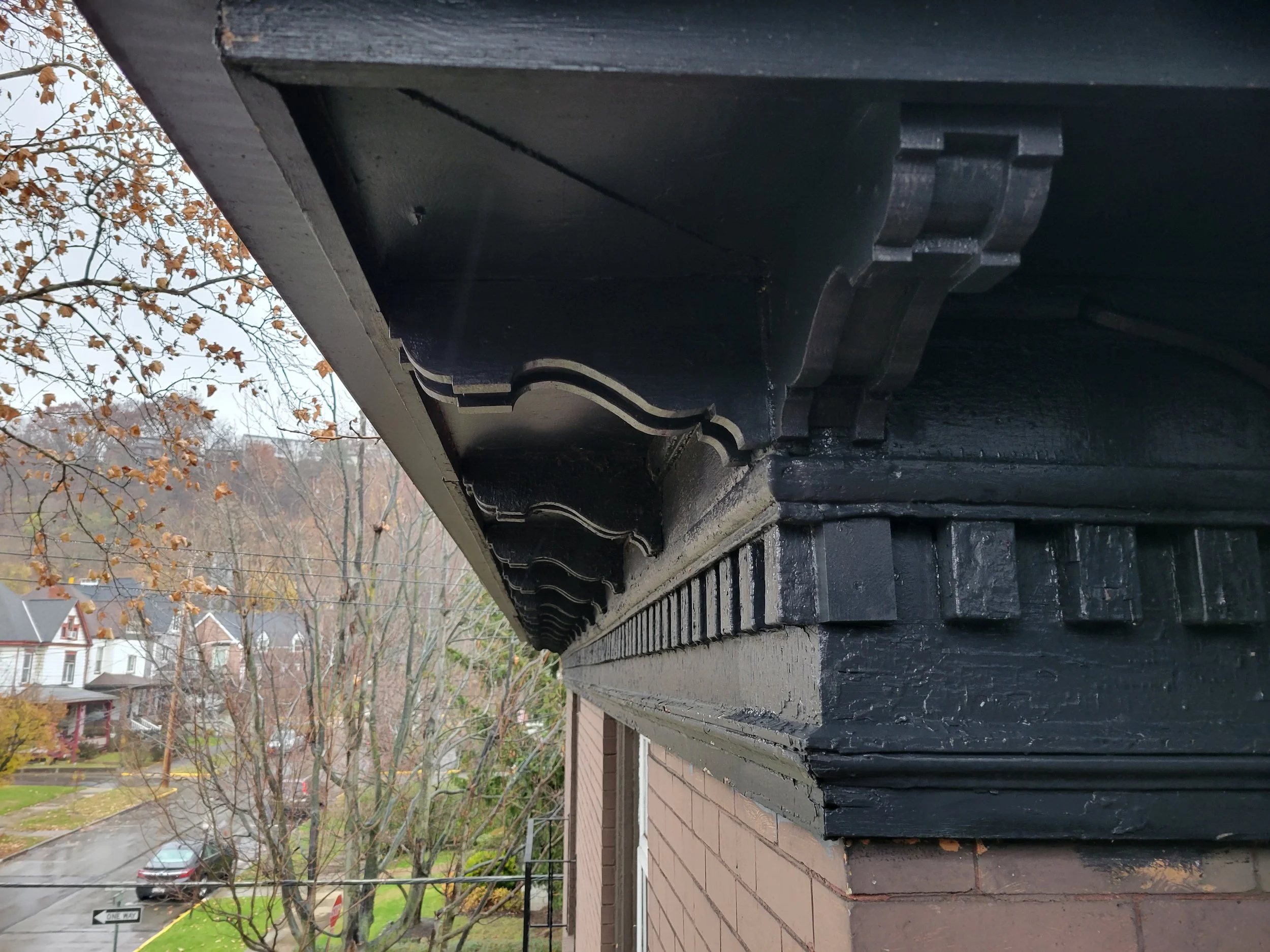 Close-up view of the black painted soffit and fascia beneath the roof edge of a brick building. In the background, a residential neighborhood with houses, trees, and a parked car on a rainy street.