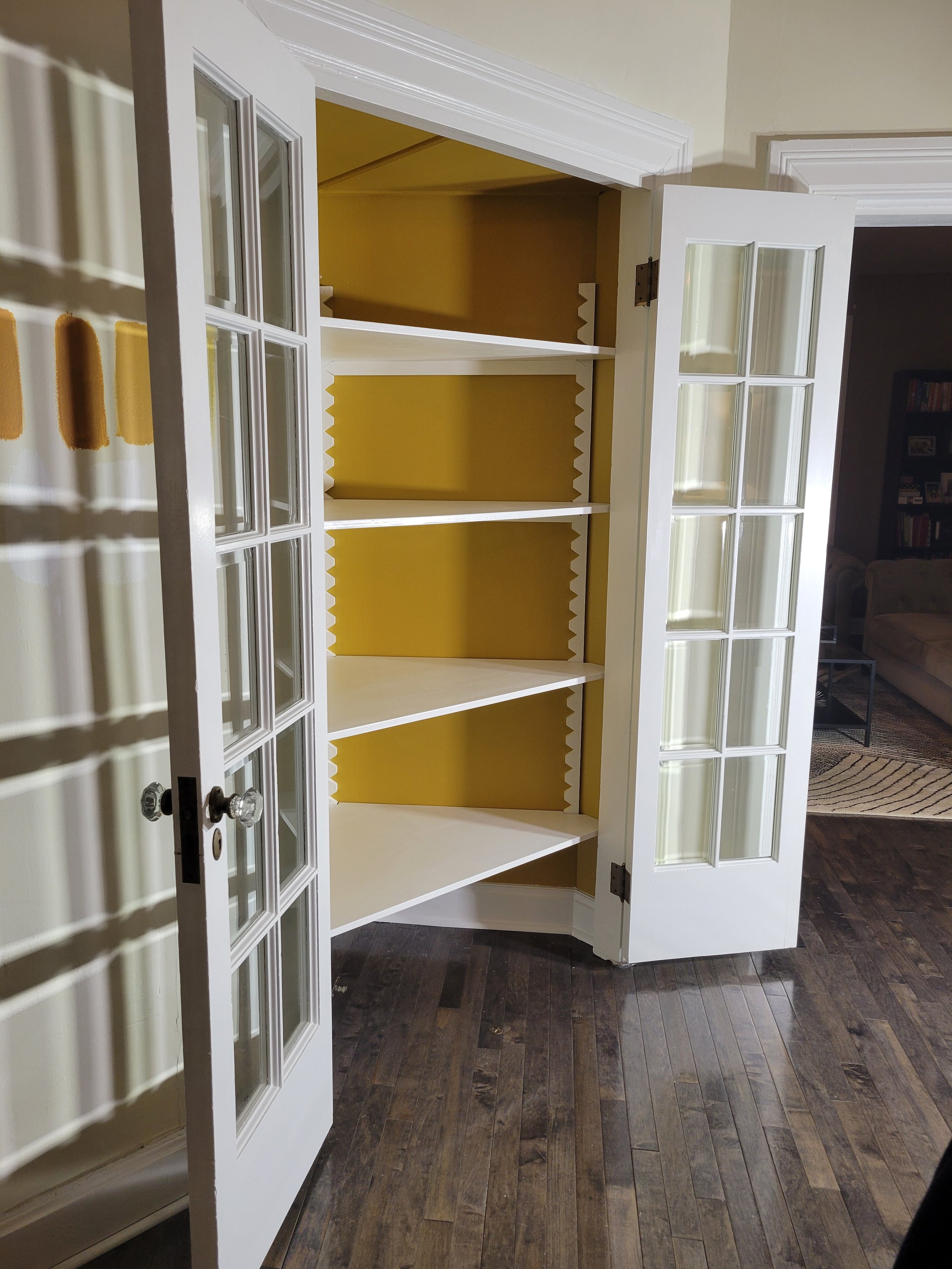 Empty white corner cabinet with glass-paneled double doors, four shelves, and yellow interior, situated in a room with dark wooden floors and a yellow wall.
