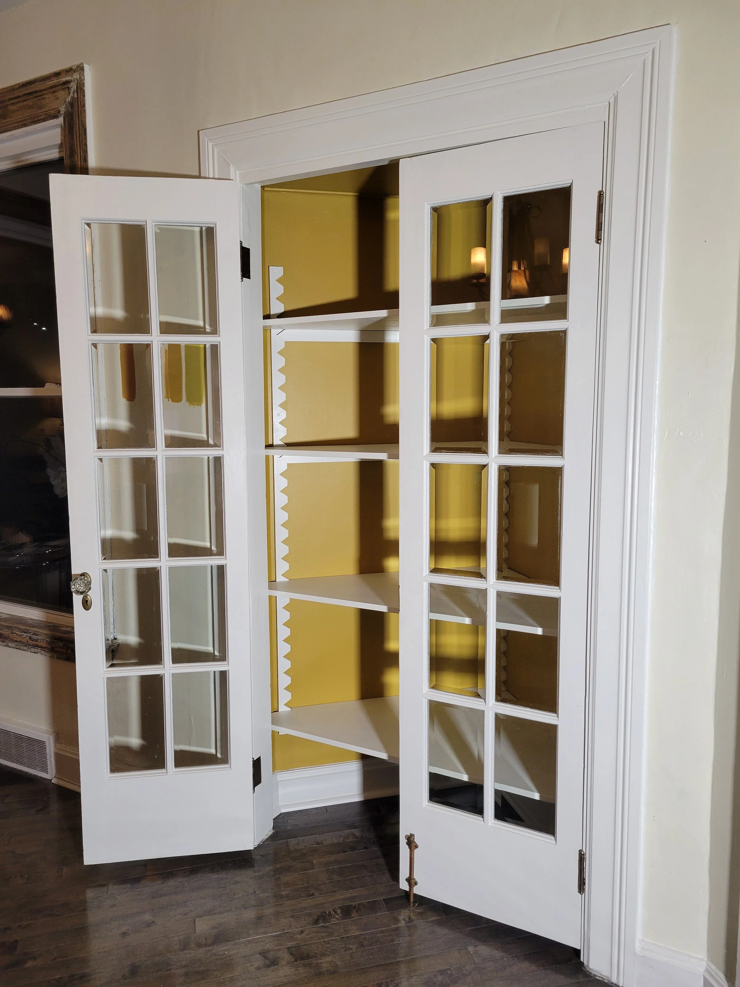 White built-in cabinet with glass-paneled doors, open to yellow walls and three shelves inside, on a dark wood floor.