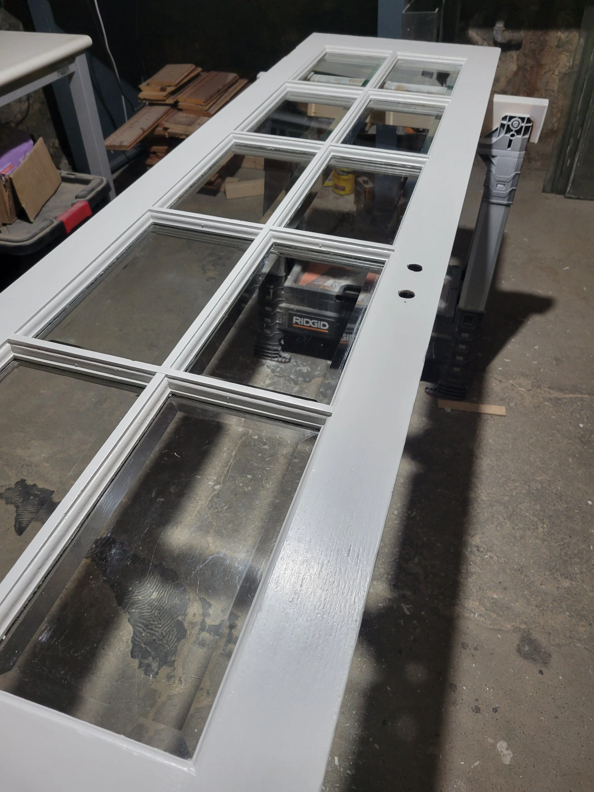 A white wooden door with nine glass panes laid on a workbench in a workshop, with various tools and materials visible in the background.
