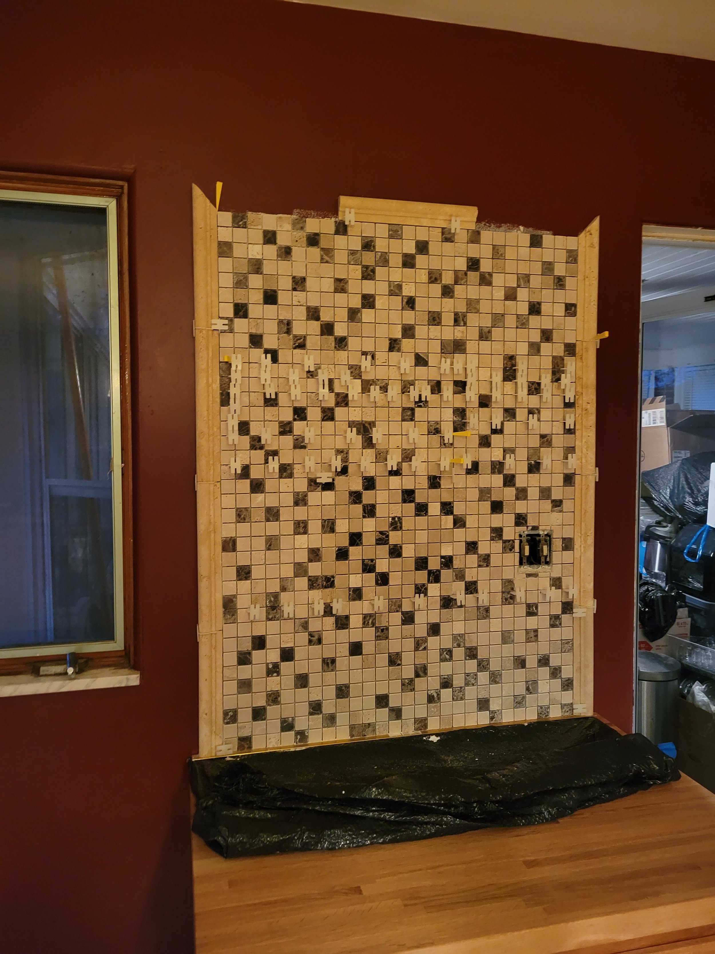Unfinished kitchen backsplash with small square tiles in shades of beige, brown, and black, framed with wood and surrounded by a red wall.