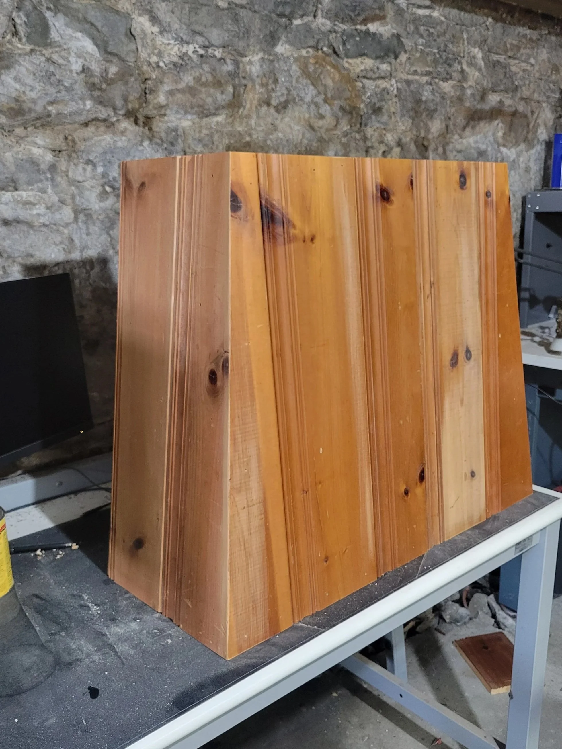 A wooden box or structure made of vertical planks with visible wood grain and knots, sitting on a table against a stone wall background.