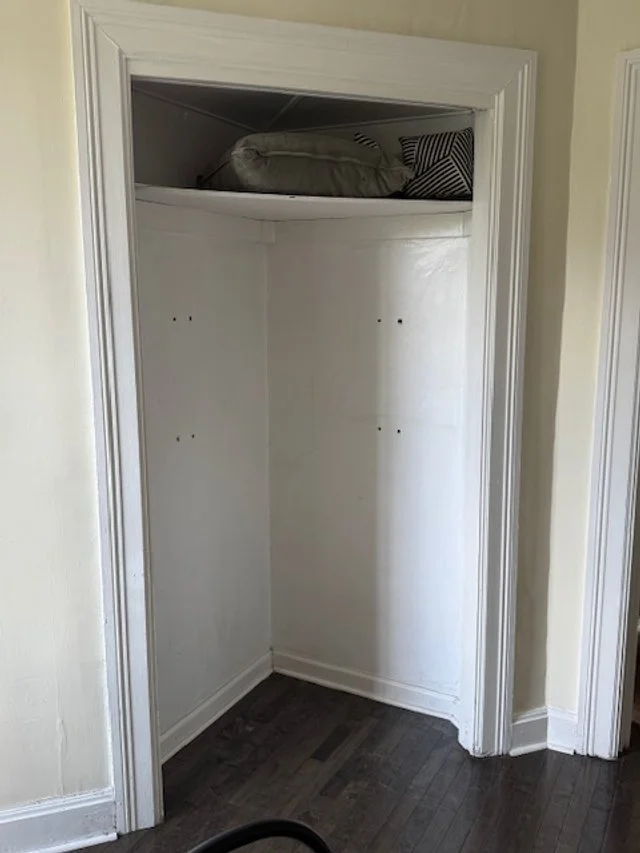 Empty built-in closet with a shelf at the top containing some pillows and bedding, with a white interior and dark hardwood floor.