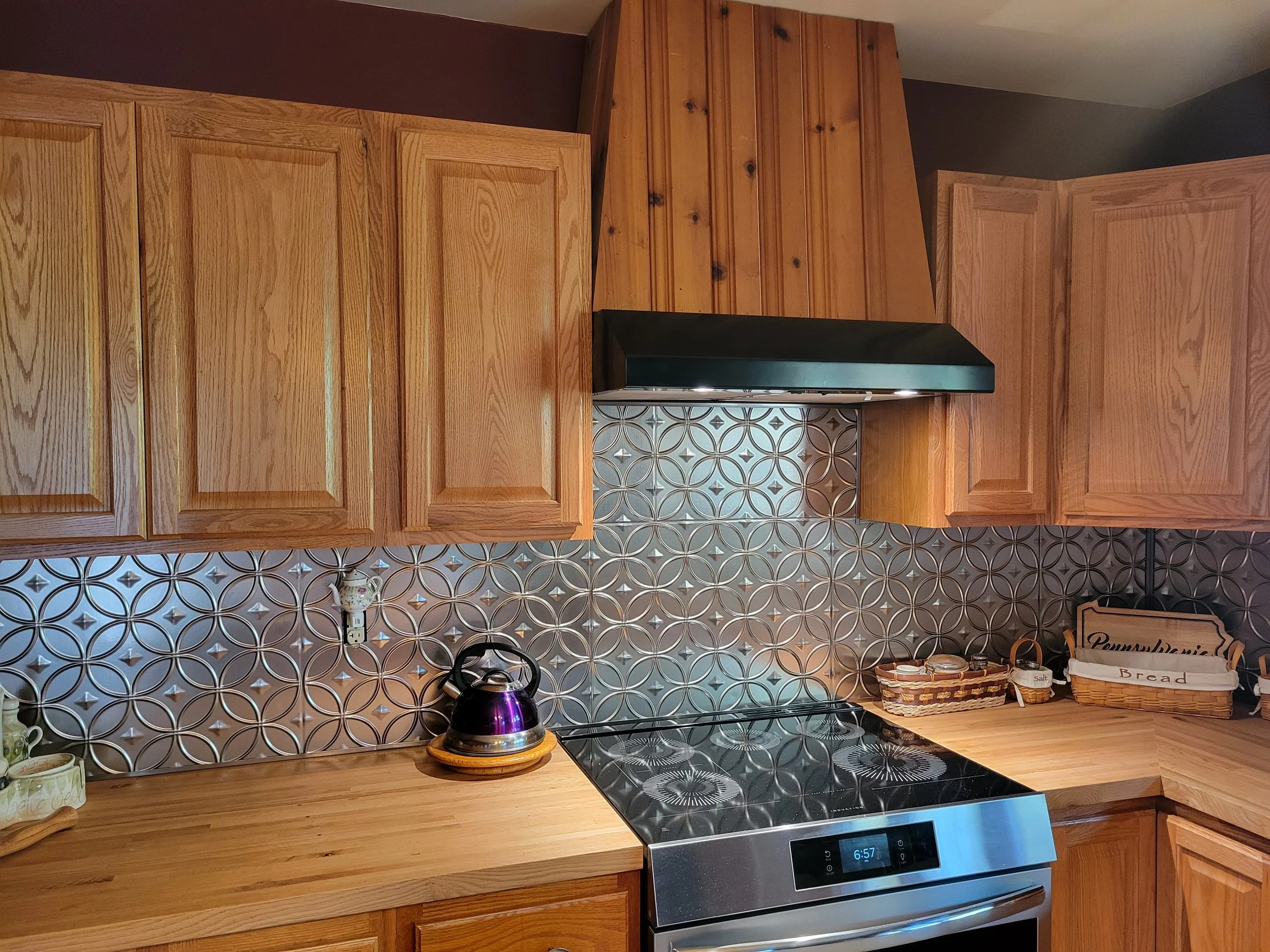 Kitchen with wooden cabinets, a black range hood, a stainless steel oven, decorative wall tiles, a purple kettle on the countertop, and baskets labeled 'Bread' and 'Pemmican'.