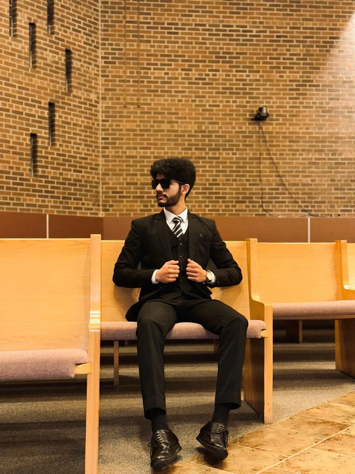 A man in a black suit with a striped tie, sunglasses, and a watch, sitting on a wooden bench in a room with brick walls and a CCTV camera.