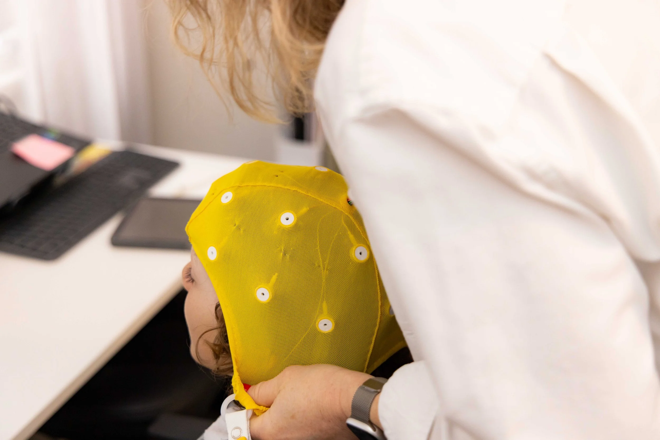 A person is holding a small child wearing a yellow hat with white buttons. The person is wearing a white shirt and has a wristband with a white strap and a gray watch. There is a black keyboard and a smartphone on the white table in the background.