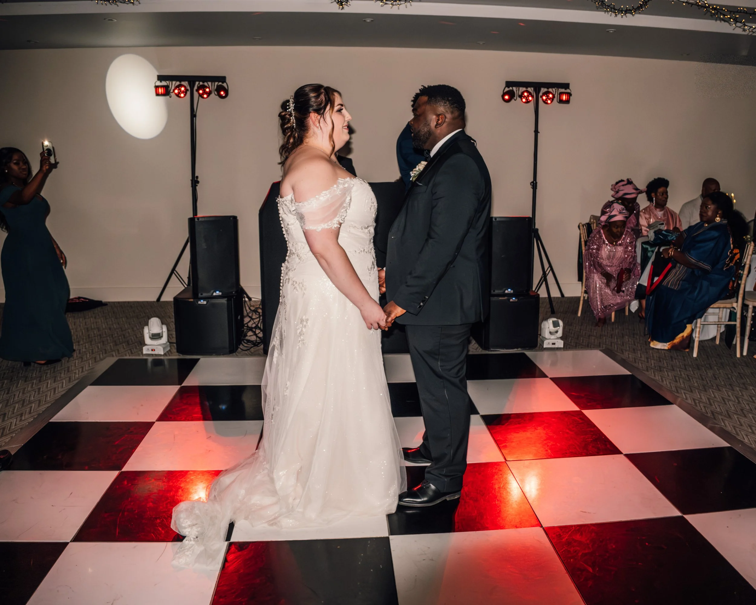 A bride and groom holding hands and smiling at each other during their wedding dance, on a decorated dance floor with guests watching in the background.