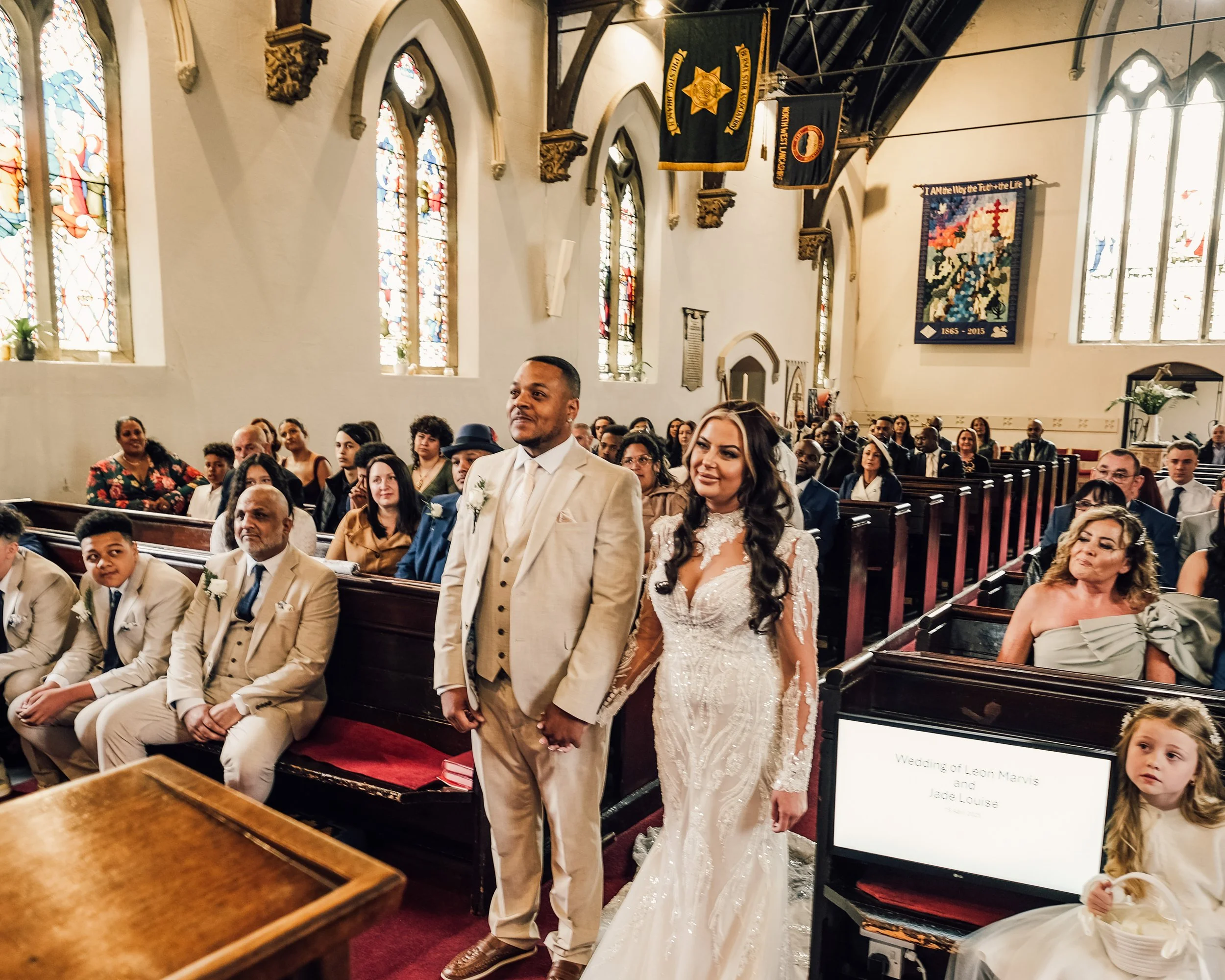 A wedding ceremony inside a church with stained glass windows and religious banners hanging from the ceiling. A bride and groom stand at the front, facing the congregation, with guests seated in pews.