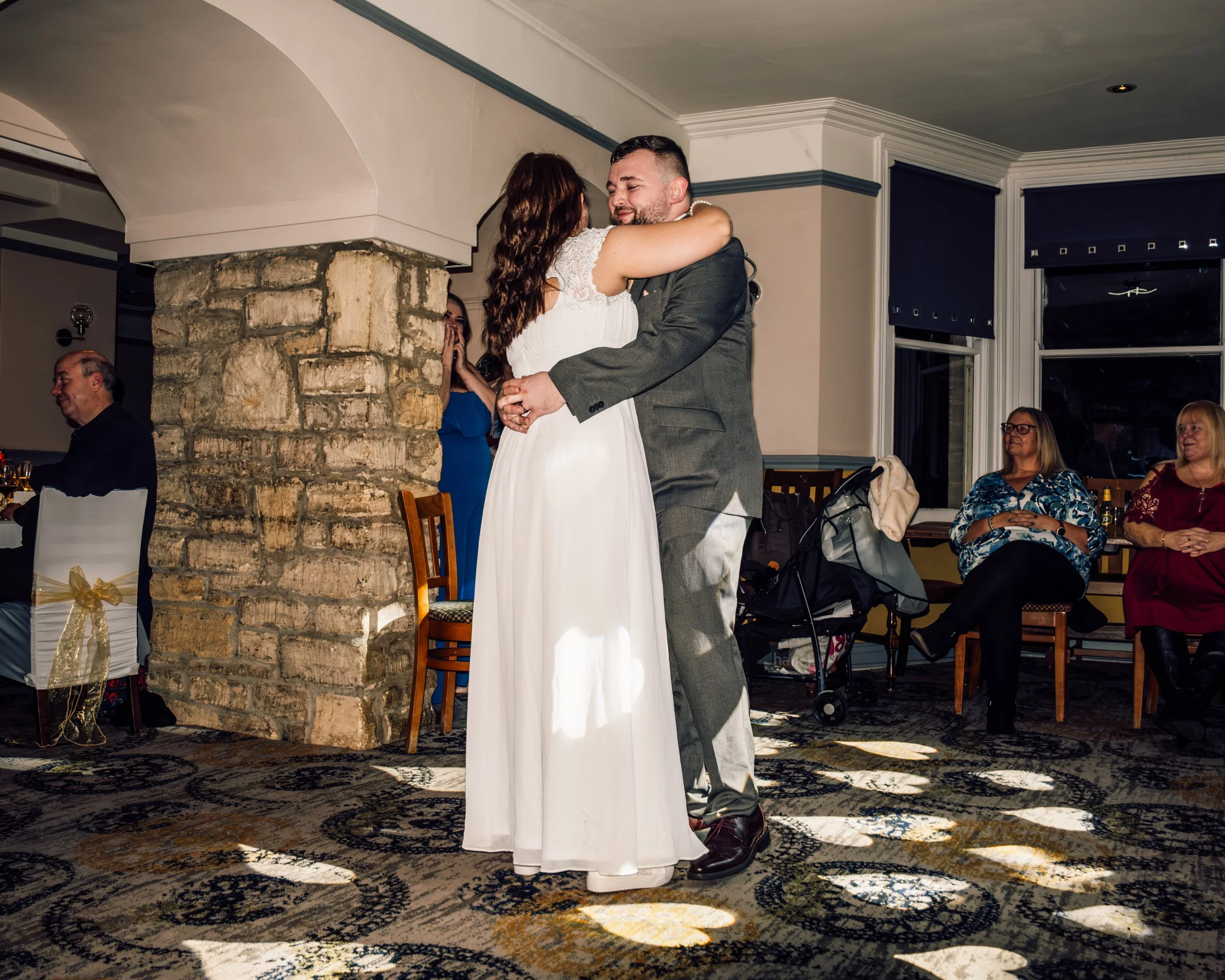 A couple dances closely at a wedding reception, with guests seated and watching in the background.
