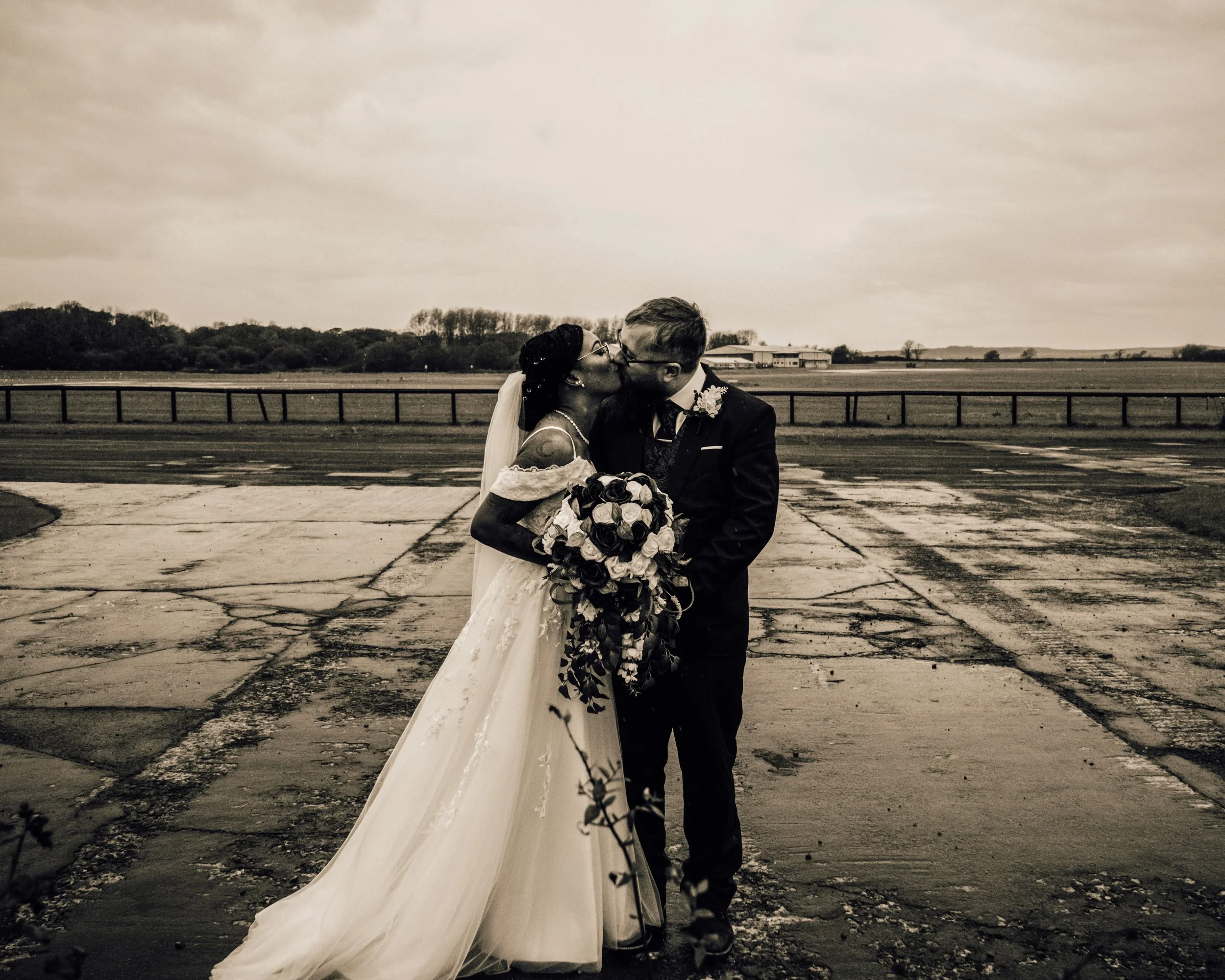 A newlywed couple kissing outdoors in black and white. The bride is holding a bouquet of flowers, and the groom is wearing a suit with a boutonniere. There is a cloudy sky and an open field in the background.