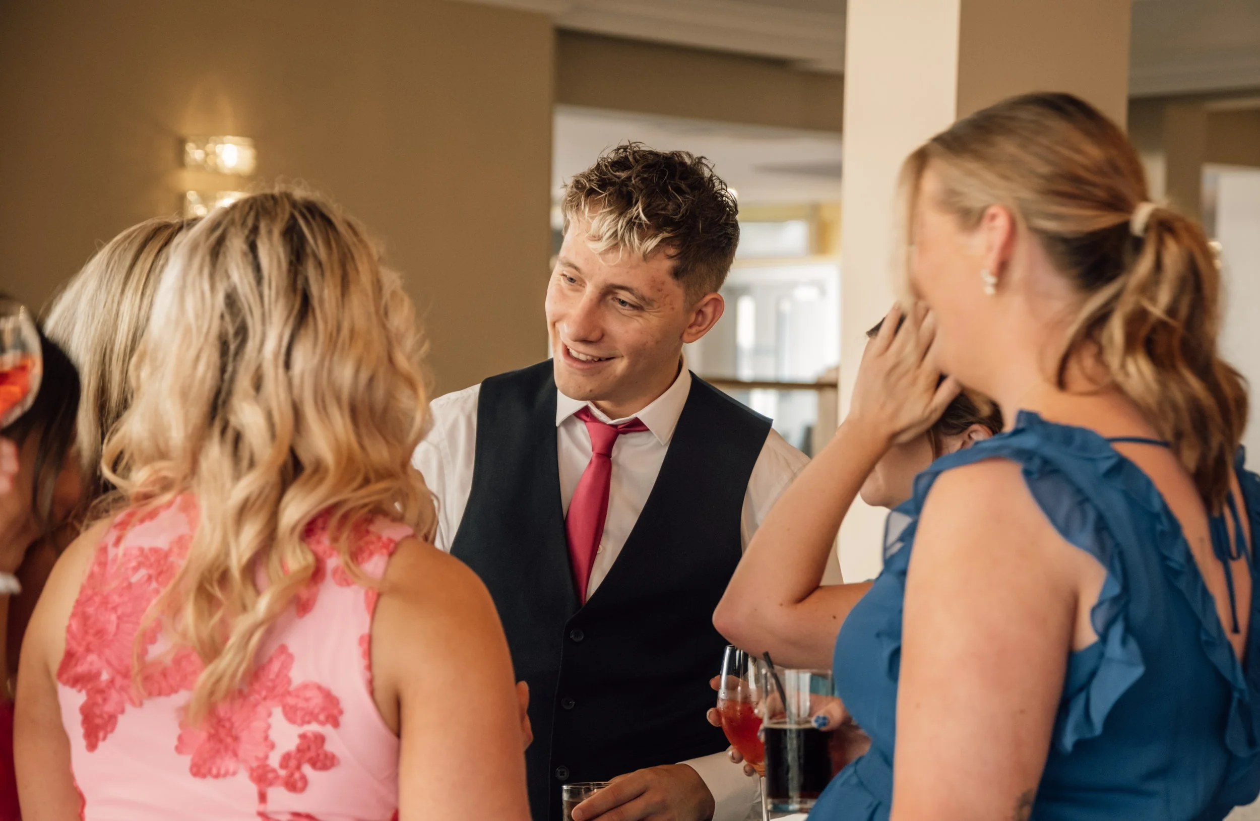 A group of people at a social event, engaged in conversation. A young man in a vest and red tie is smiling at the women around him. One woman in a pink dress and another in a blue dress are talking, holding drinks.