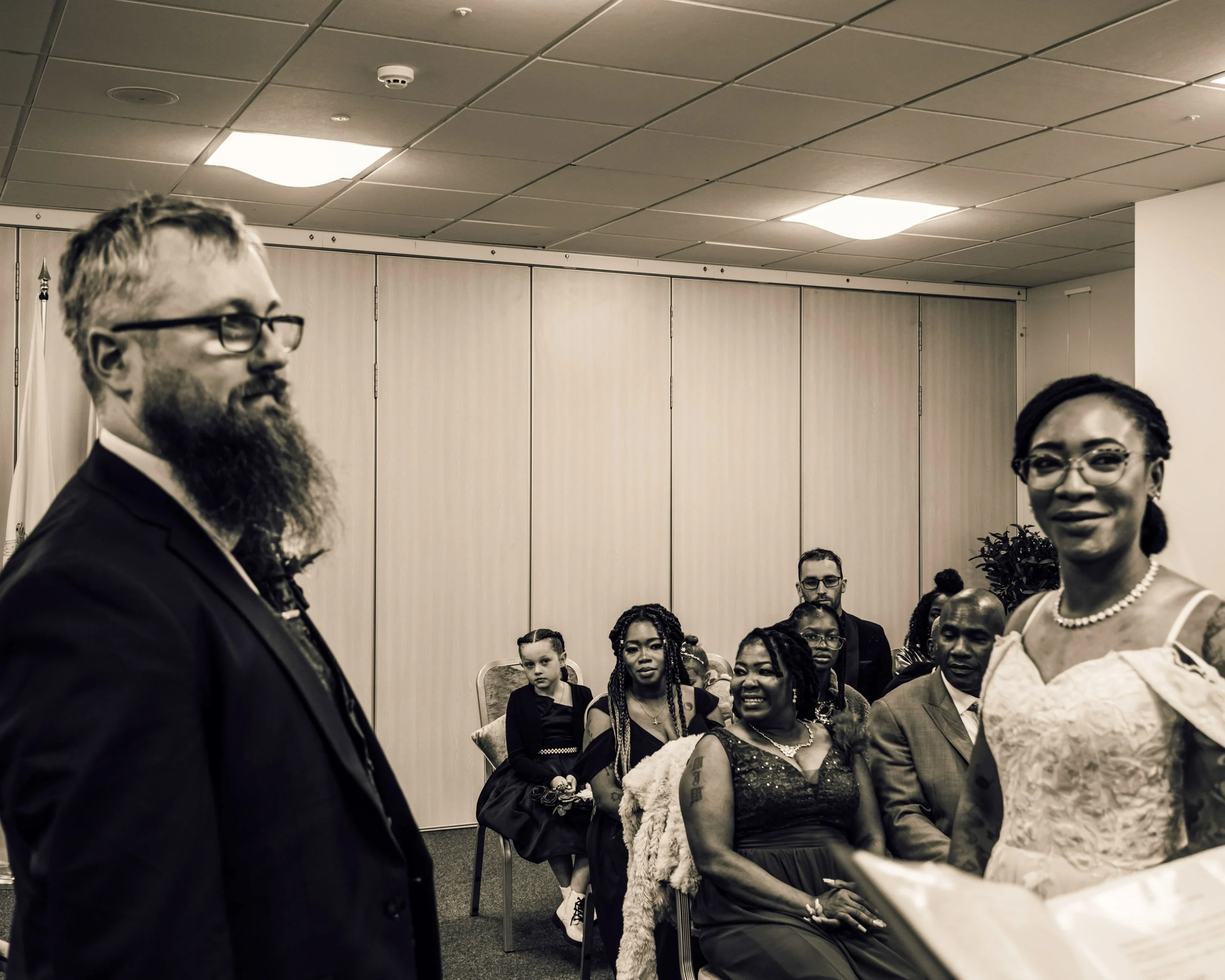 A group of people at a formal event or gathering, seated and standing in a room with a drop ceiling and soft lighting. The attendees are dressed in business or formal attire, including dresses, suits, and jewelry. The focus is on a woman with glasses