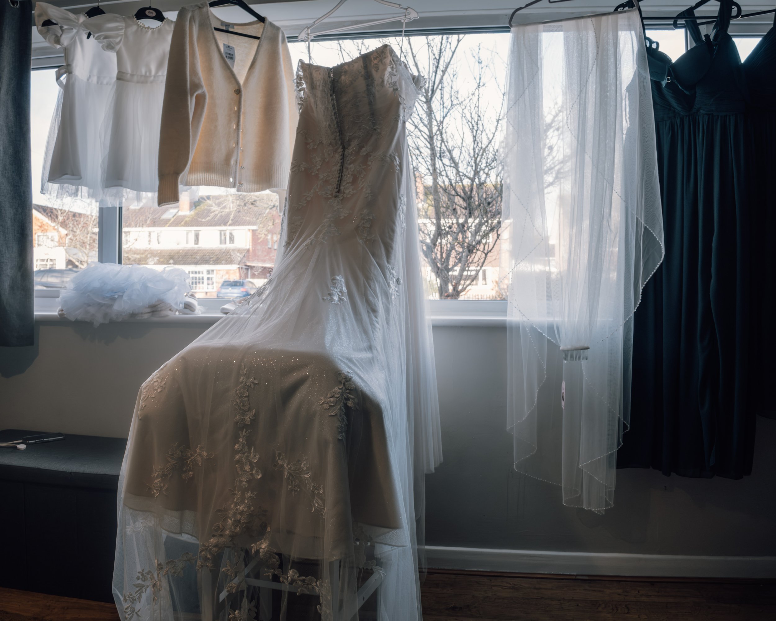 A wedding dress hanging in a window, surrounded by sheer curtains and other pieces of clothing, with trees and buildings visible outside.