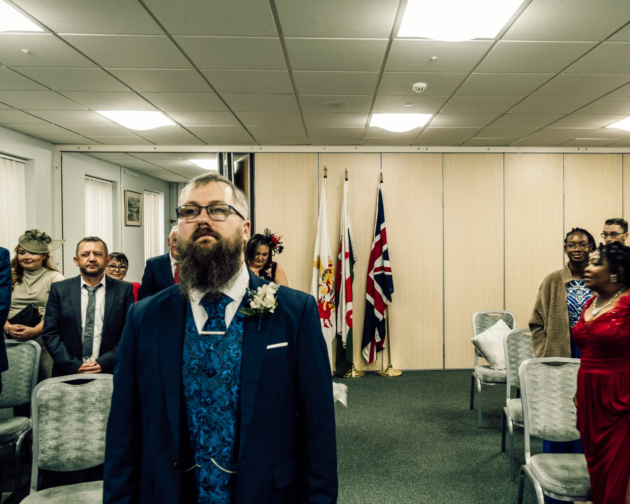 People attending a formal event or ceremony, some dressed in colorful attire, standing in a room with flags and chairs, with a man in the foreground wearing glasses and a suit.