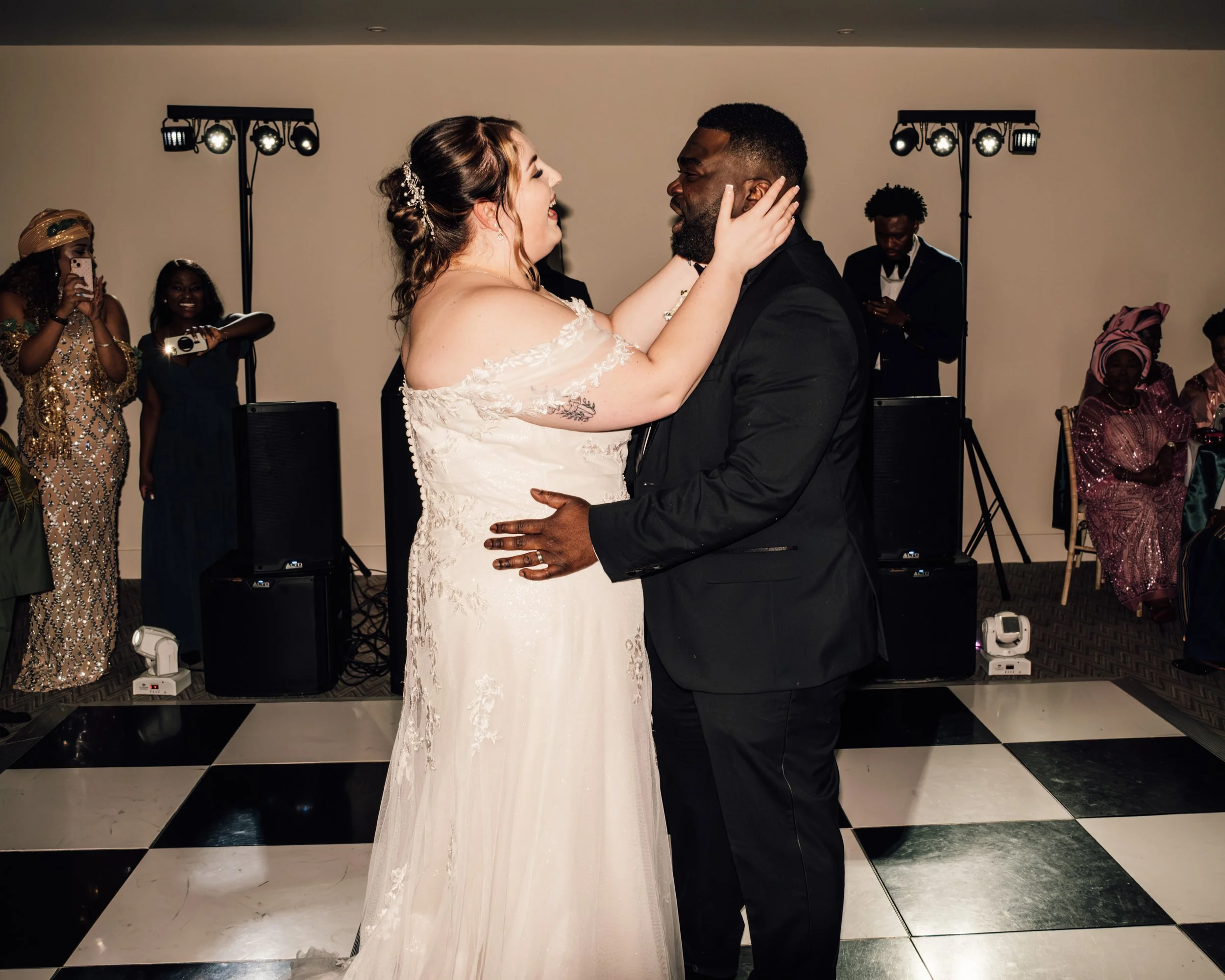 A bride and groom dancing at their wedding reception, surrounded by guests in formal attire, with stage lights overhead and a black-and-white checkered dance floor.