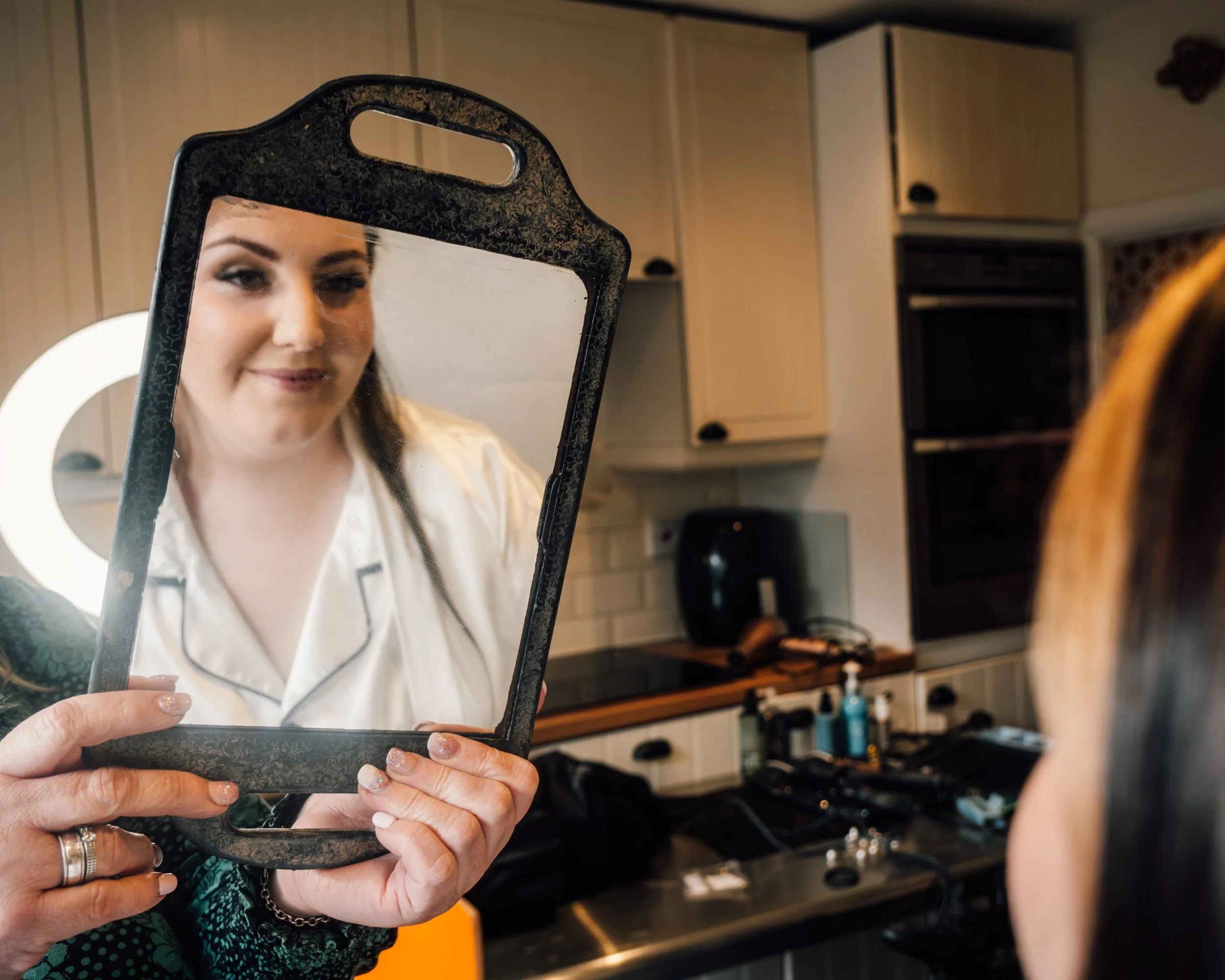 Person with manicured fingers holding a small mirror reflecting her face, which shows a friendly expression, in a room resembling a salon or kitchen.