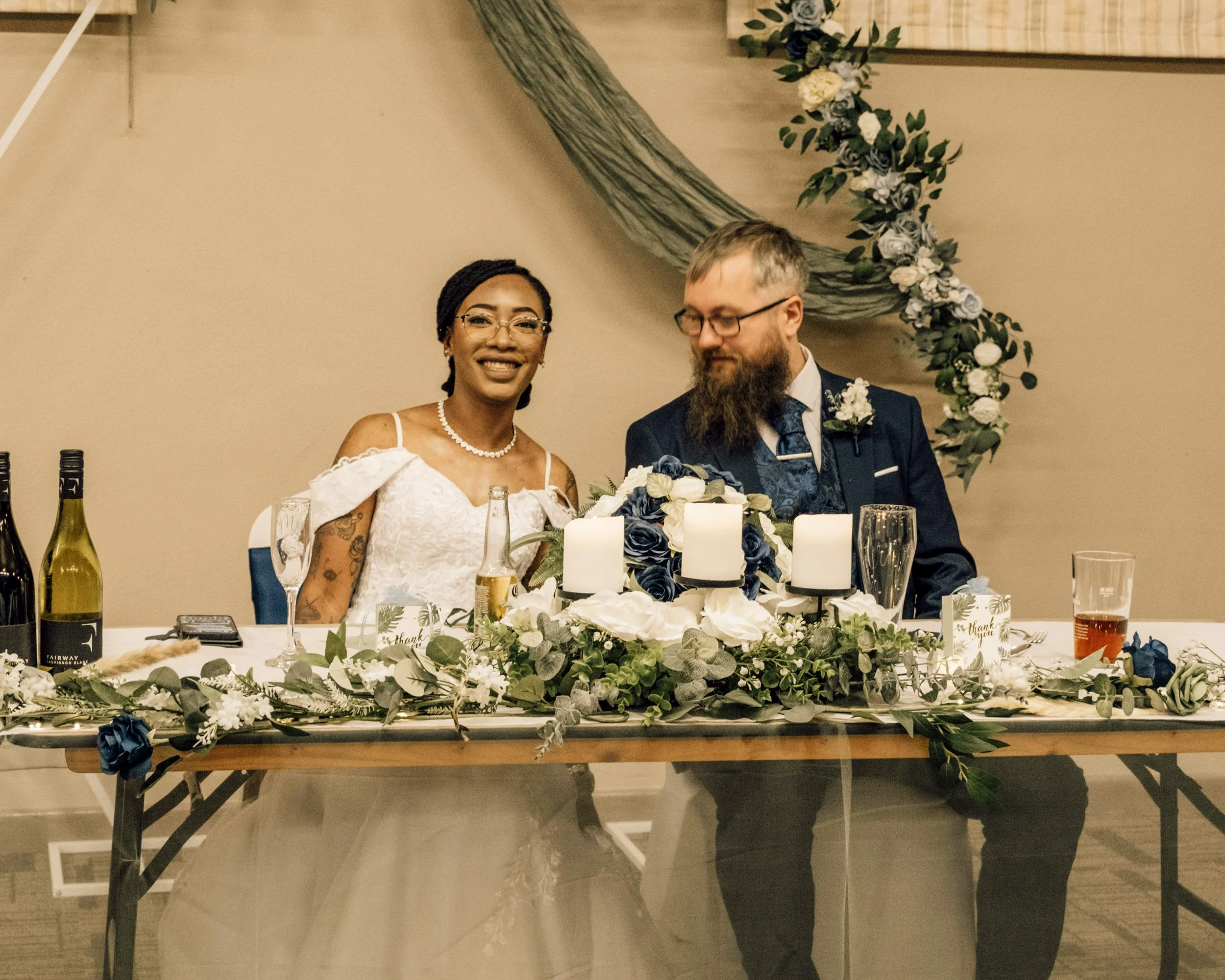 A bride and groom sitting at a decorated wedding reception table with floral arrangements and candles, smiling and looking at each other. The bride is wearing a white wedding dress and glasses, and the groom is in a dark suit with glasses and a beard