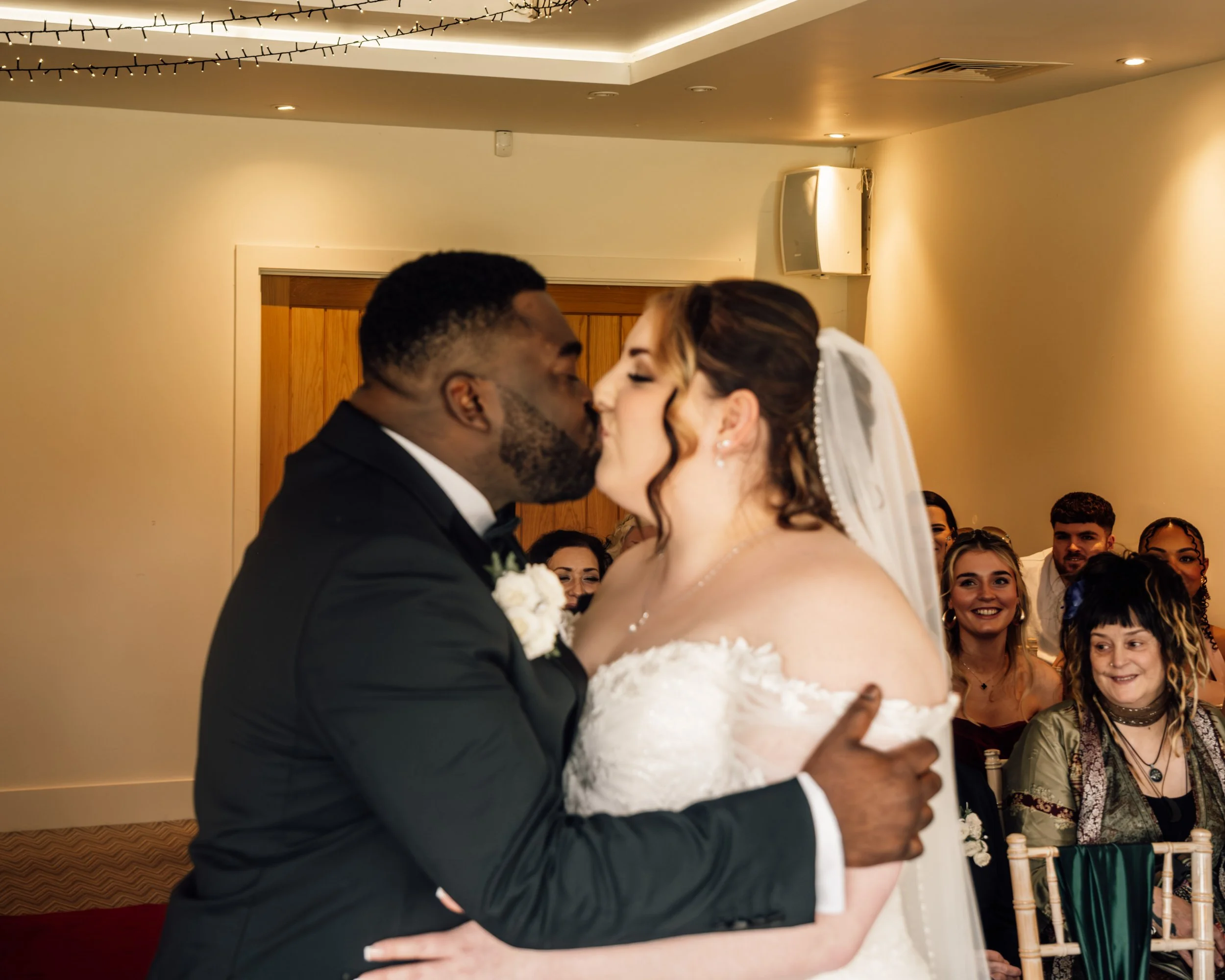 A bride and groom share a kiss during their wedding ceremony, surrounded by seated guests in a decorated indoor venue.