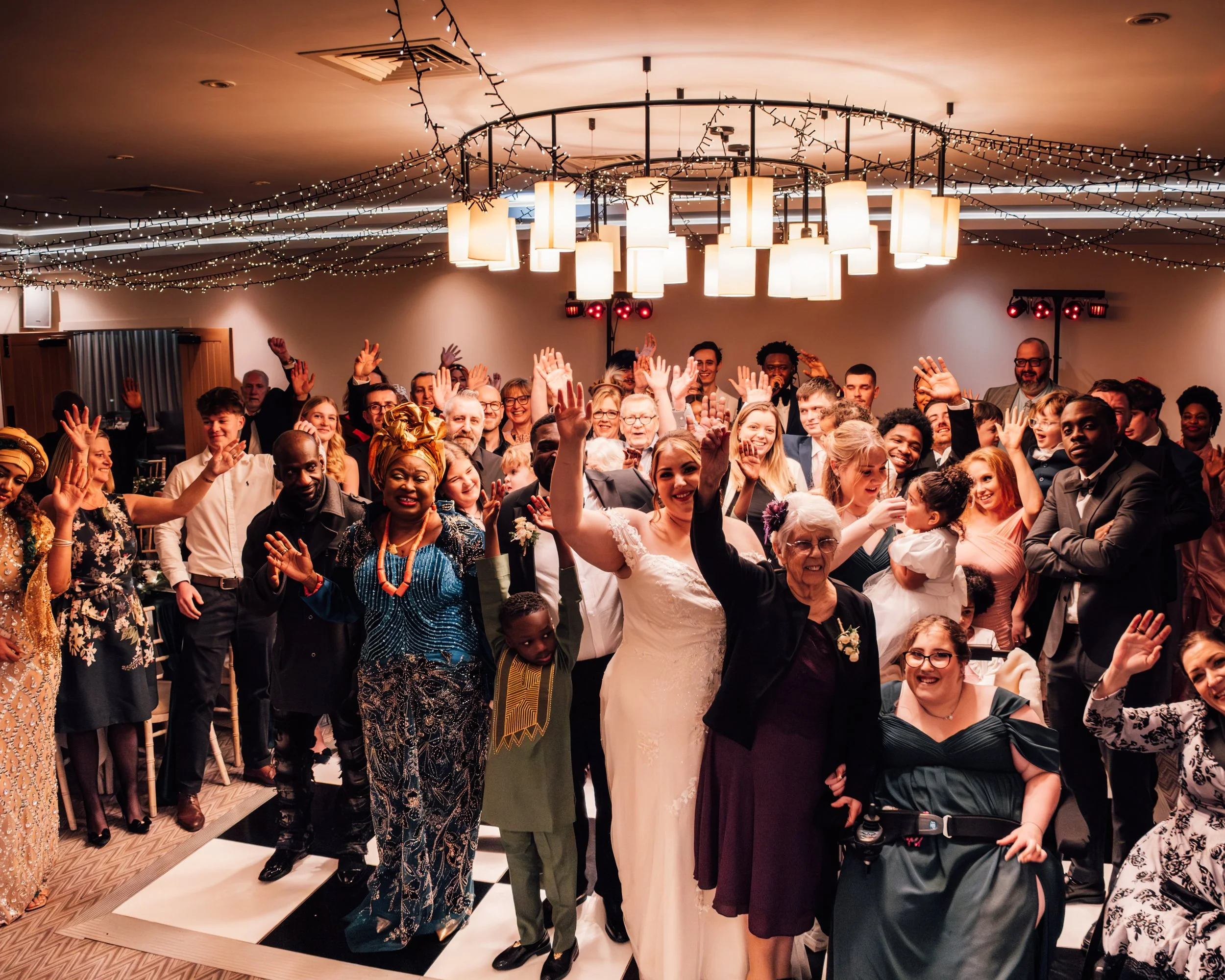 Crowd of diverse people at a celebration or wedding, many with raised hands, in a decorated indoor venue with string lights and modern lighting fixtures.