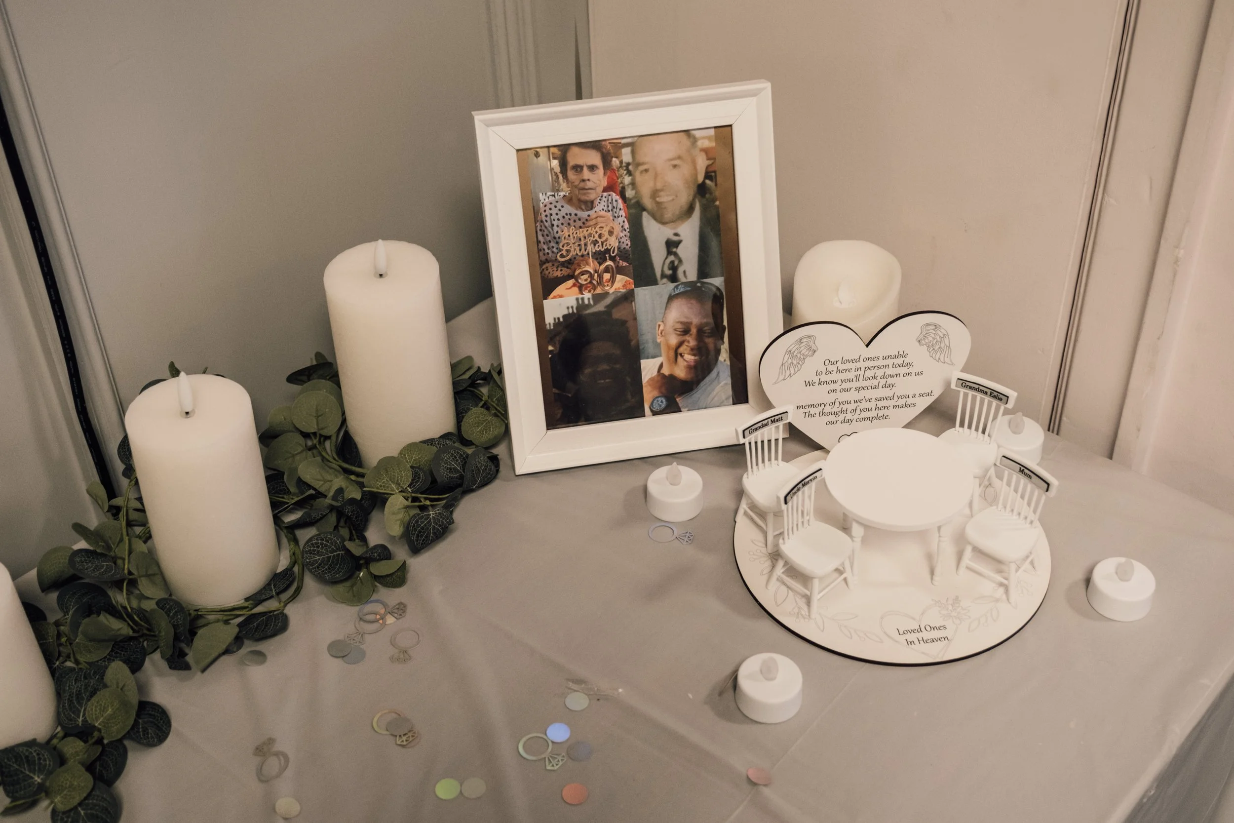 Memorial display with framed photos of loved ones, surrounded by white candles, decorative leaves, and a round table with miniature chairs, honoring deceased family members.