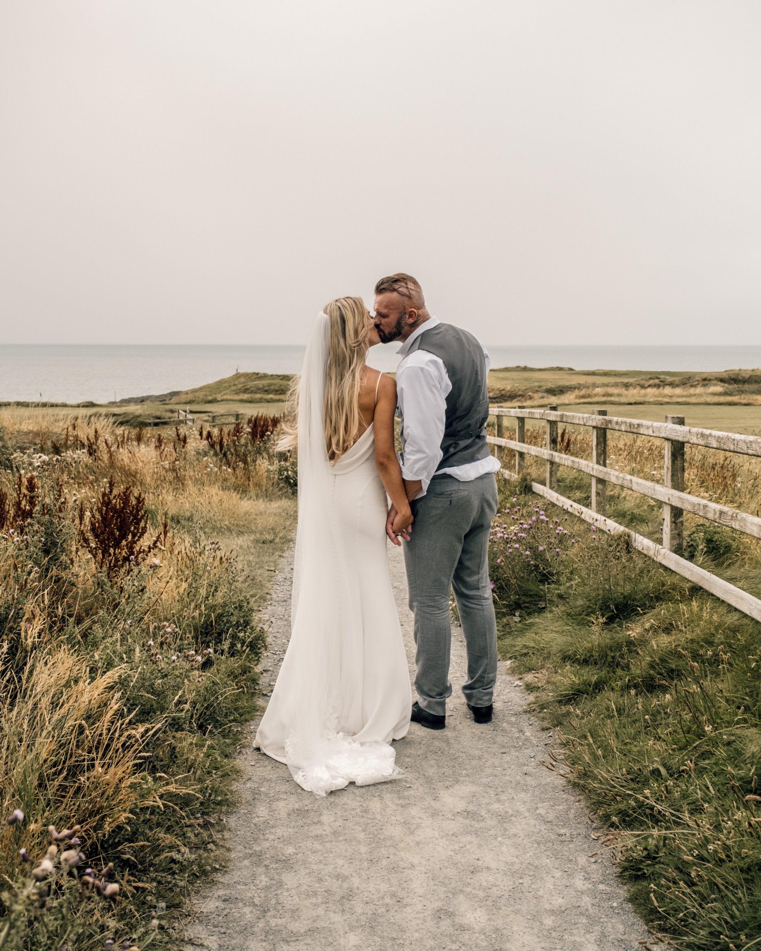 A couple dressed in wedding attire sharing a kiss on a dirt path by the sea, with grassy fields and a wooden fence in the background.