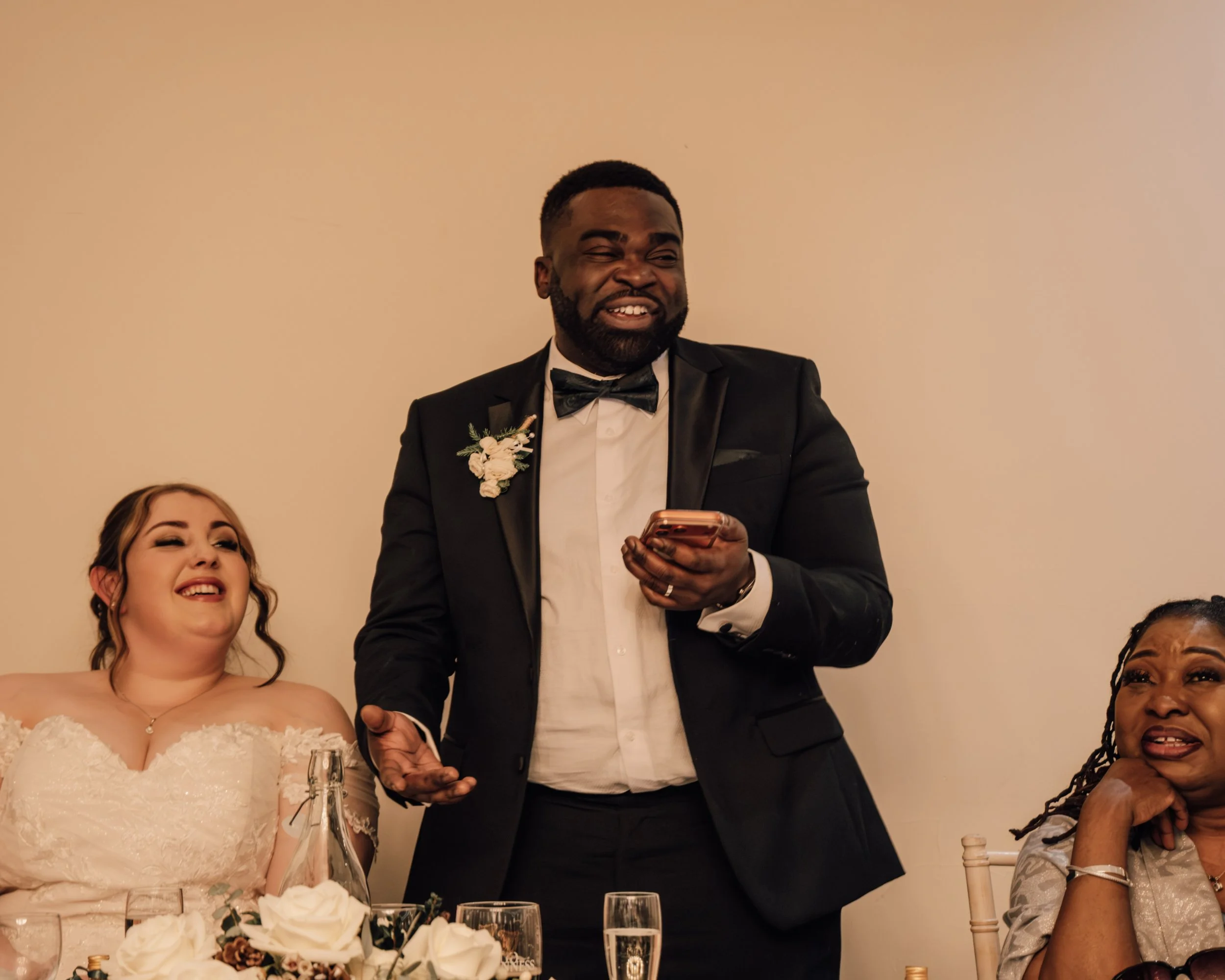 A group of people celebrating at a formal event, with a man in a tuxedo holding a phone and smiling, and a woman in a white lace dress laughing, surrounded by floral arrangements and glasses on a table.