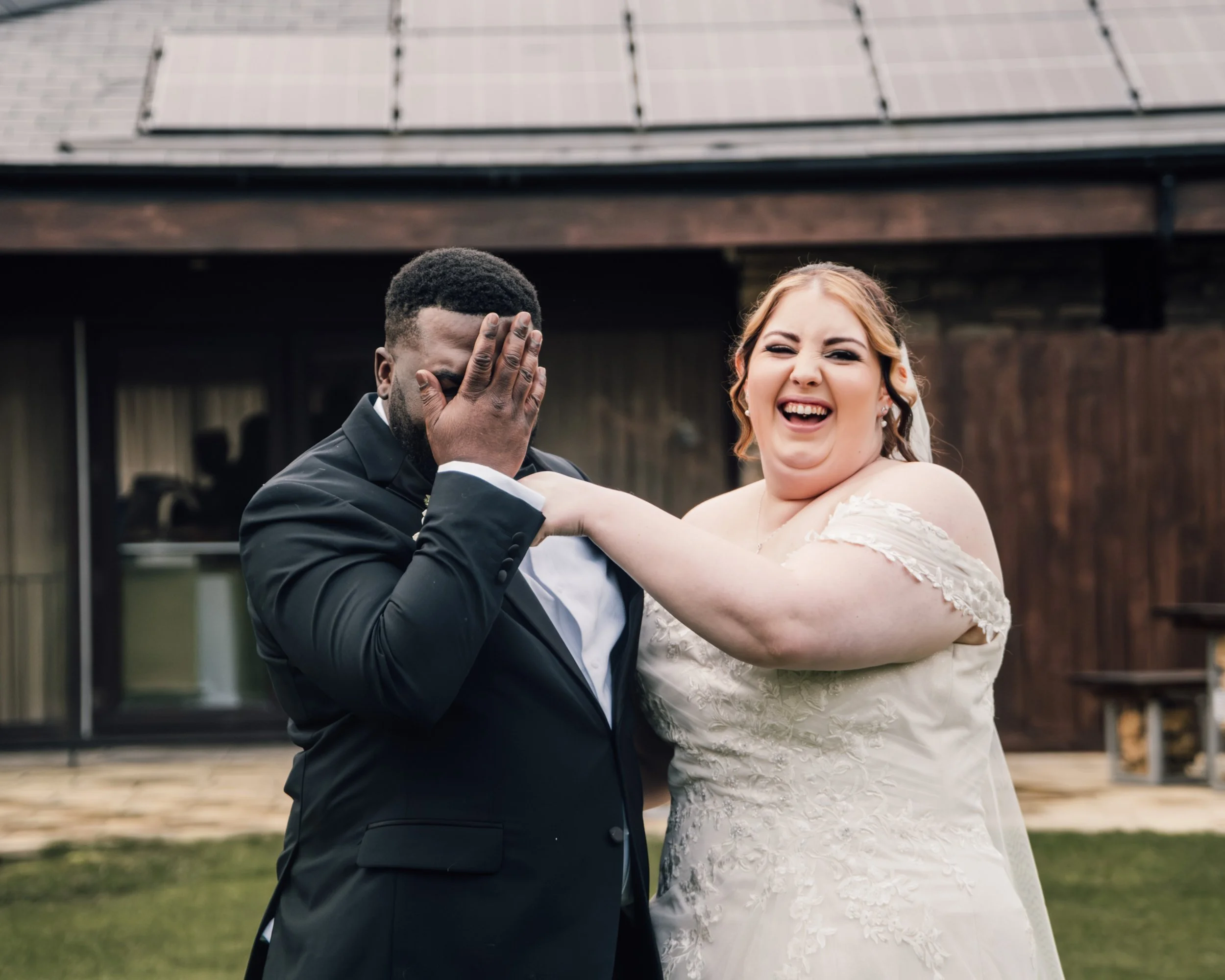 A smiling bride and groom dancing outdoors at their wedding. The groom is covering his face with his hand, and the bride is laughing with her arm around him.