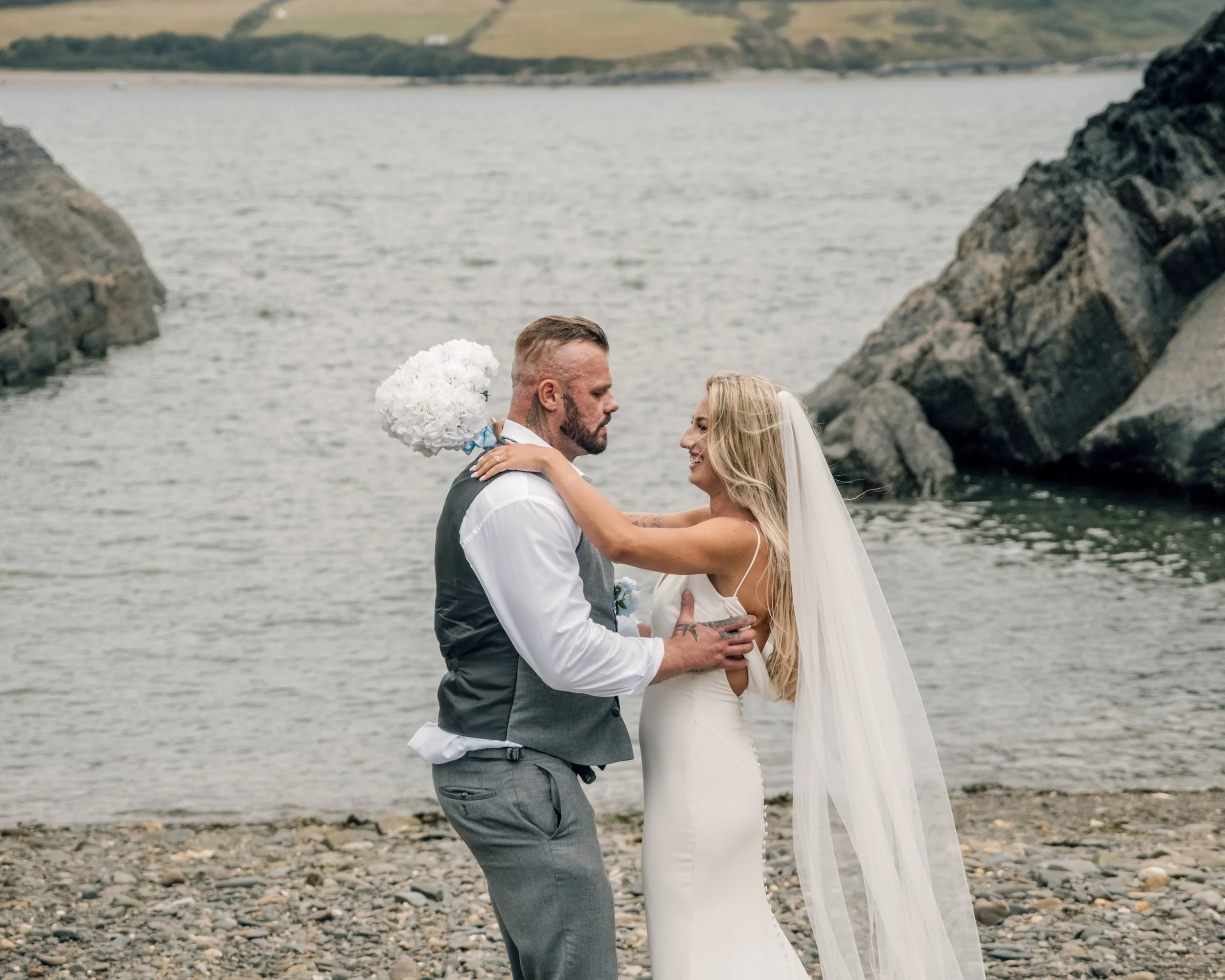 A couple in wedding attire standing on a rocky beach, embracing each other with a body of water and green hills in the background.