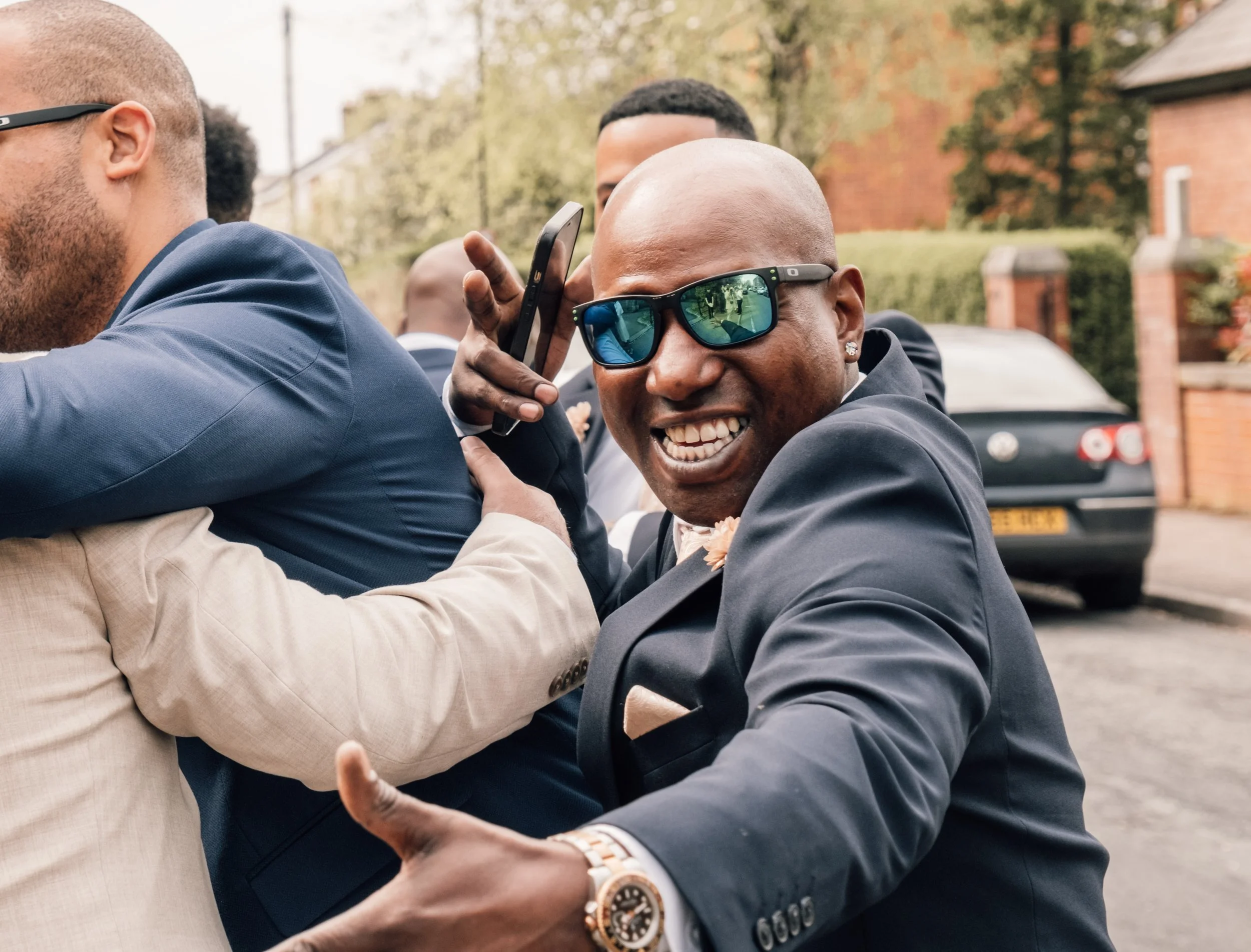 Several well-dressed men, some wearing sunglasses, are engaged in a joyful altercation outdoors, smiling and appearing to enjoy each other's company.