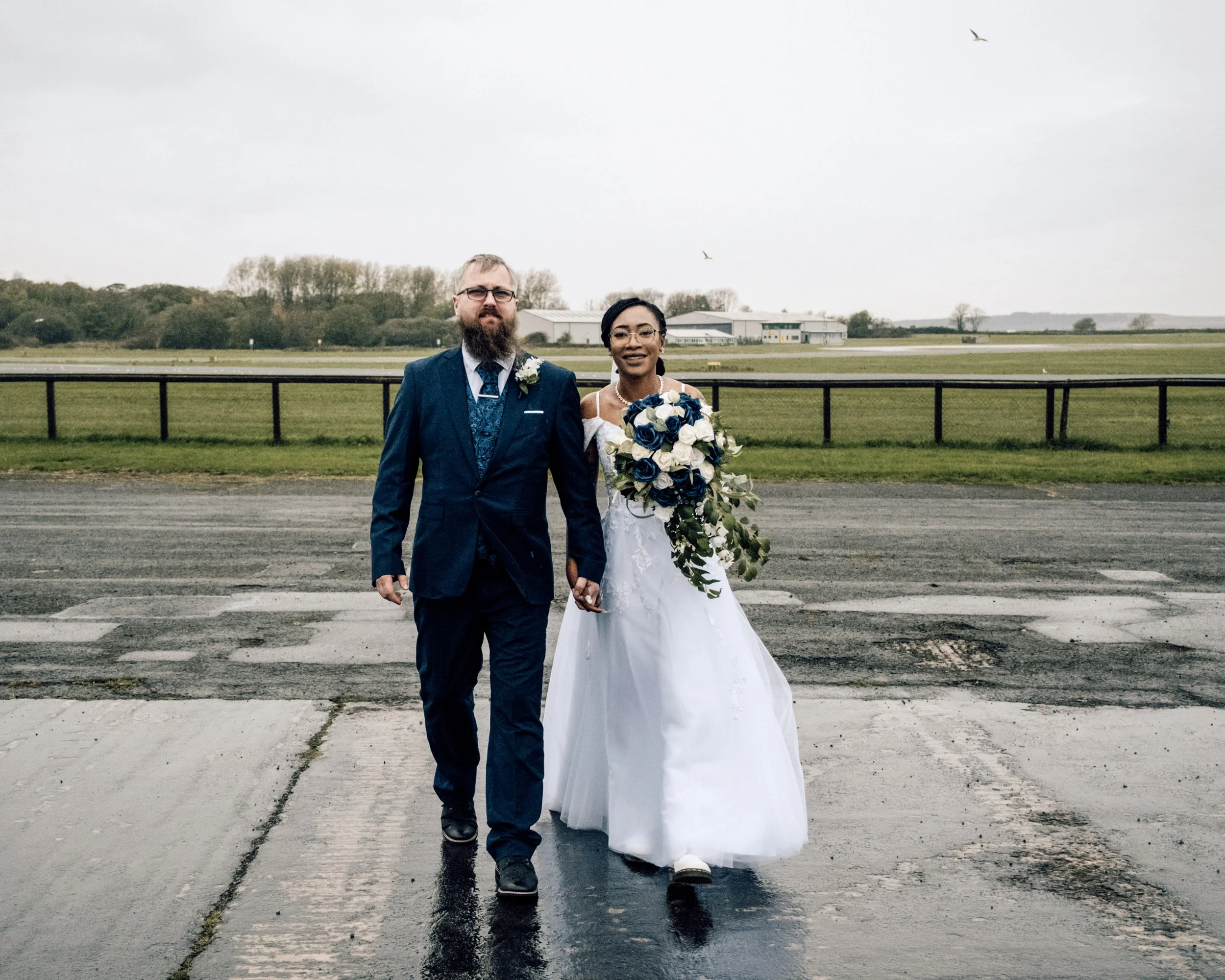 A man in a blue suit and a woman in a white wedding dress holding a bouquet of white and blue roses walking together outdoors on a cloudy day.