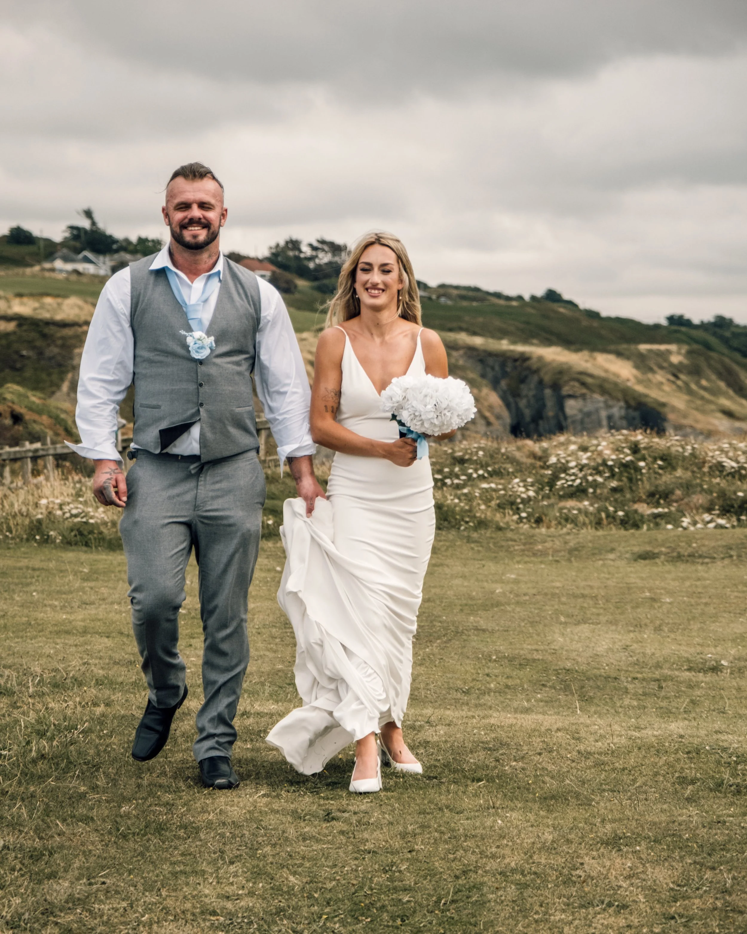A bride and groom walking outdoors on a cloudy day, the bride holding a bouquet of white flowers, both smiling, with green hills and houses in the background.