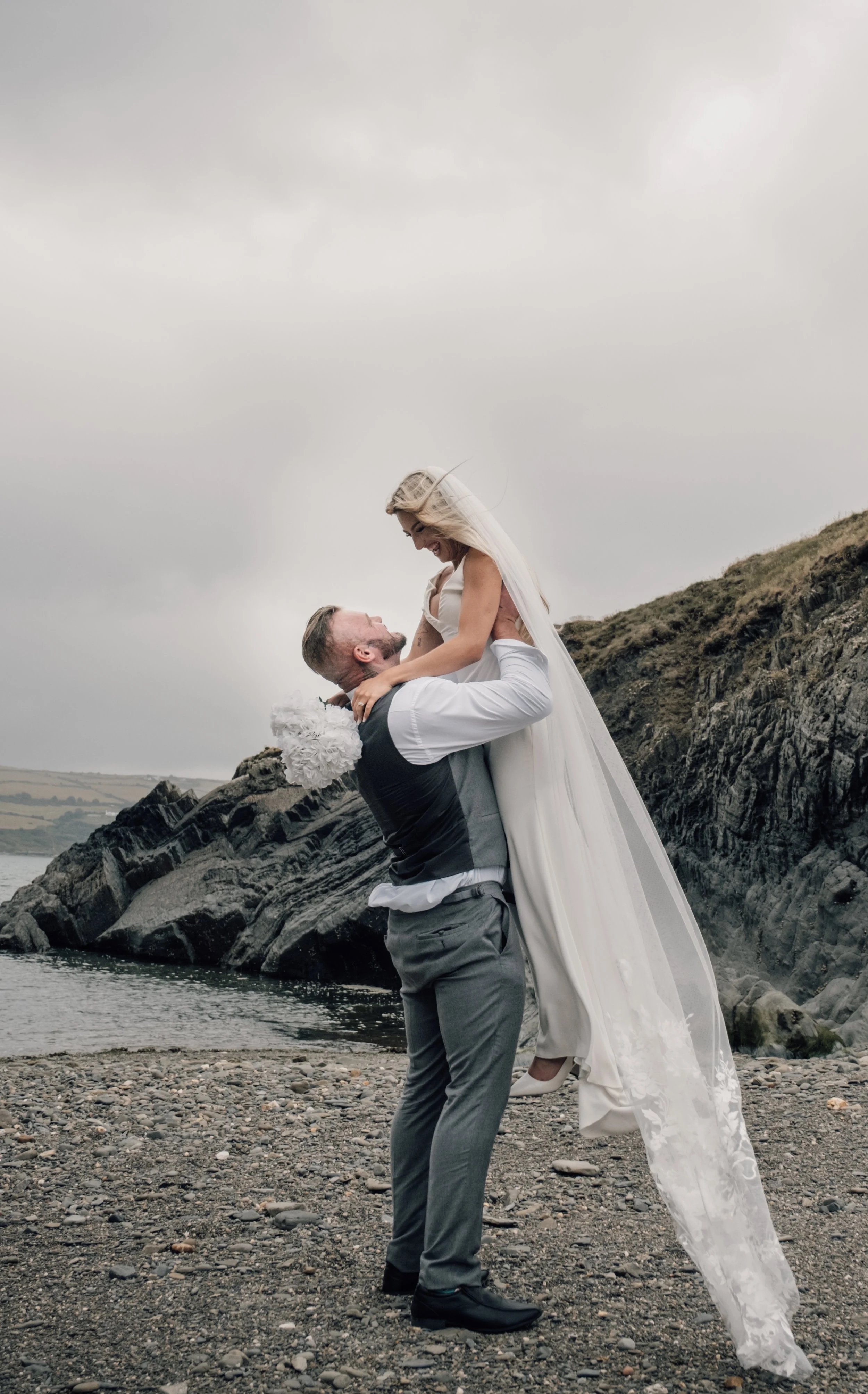 A man in a gray vest and pants lifting a woman in a white wedding dress and veil in his arms on a rocky beach with cliffs and cloudy sky in the background, celebrating their wedding.