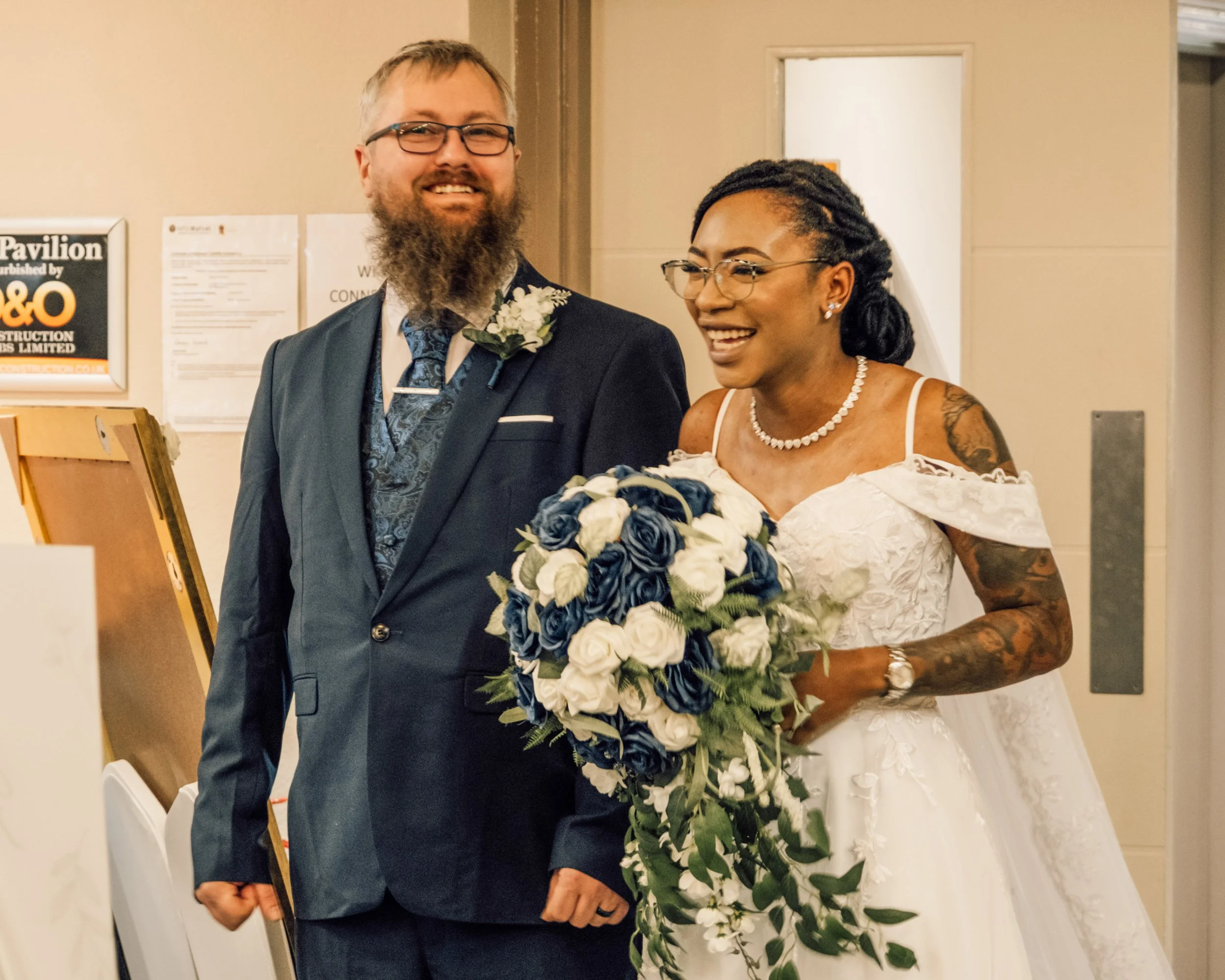 A man in a suit with glasses and a beard smiling next to a woman in a white wedding dress with glasses and tattoos holding a bouquet of white and blue flowers.
