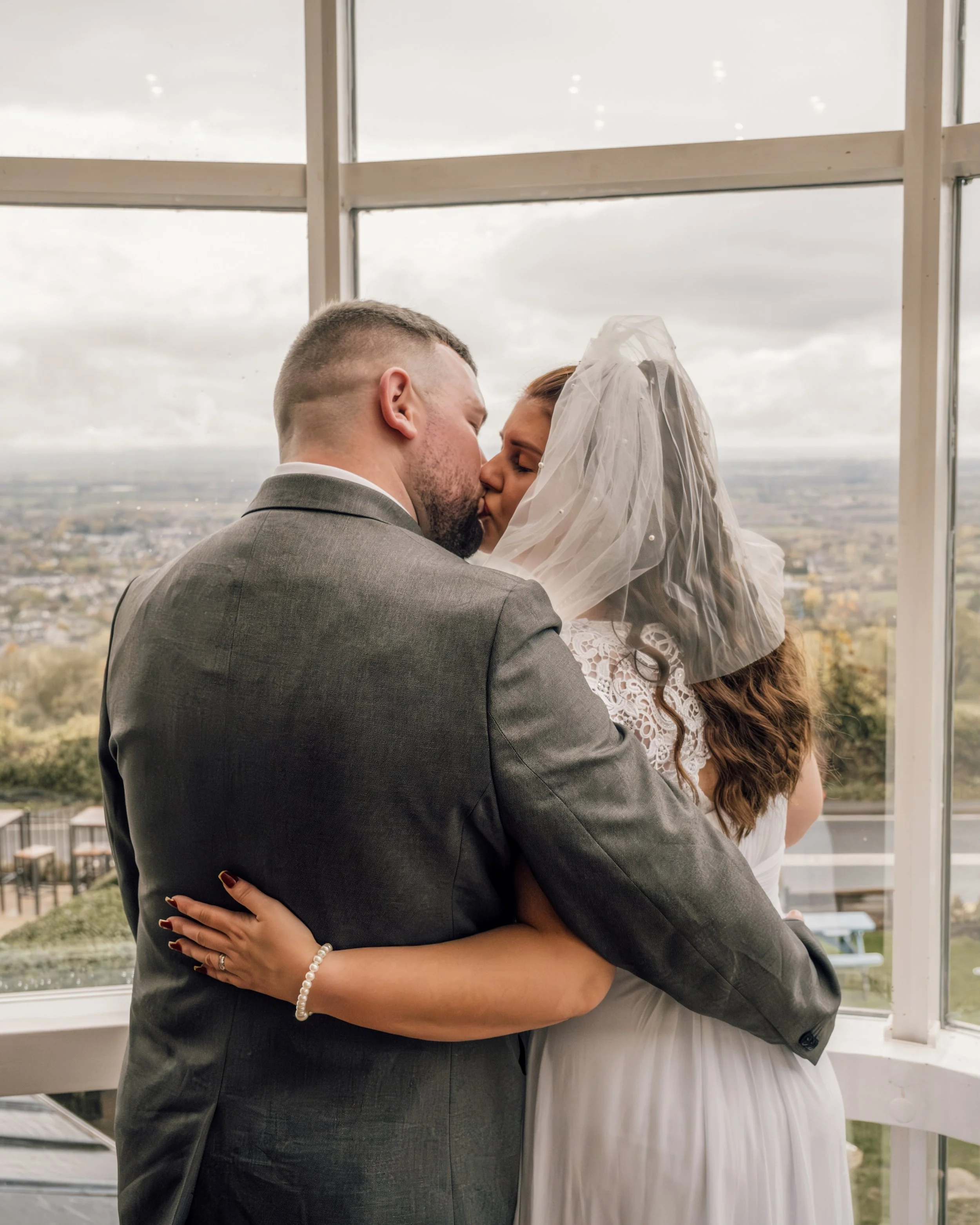 A newlywed couple sharing a kiss indoors with a large window behind them, revealing a cityscape and cloudy sky.
