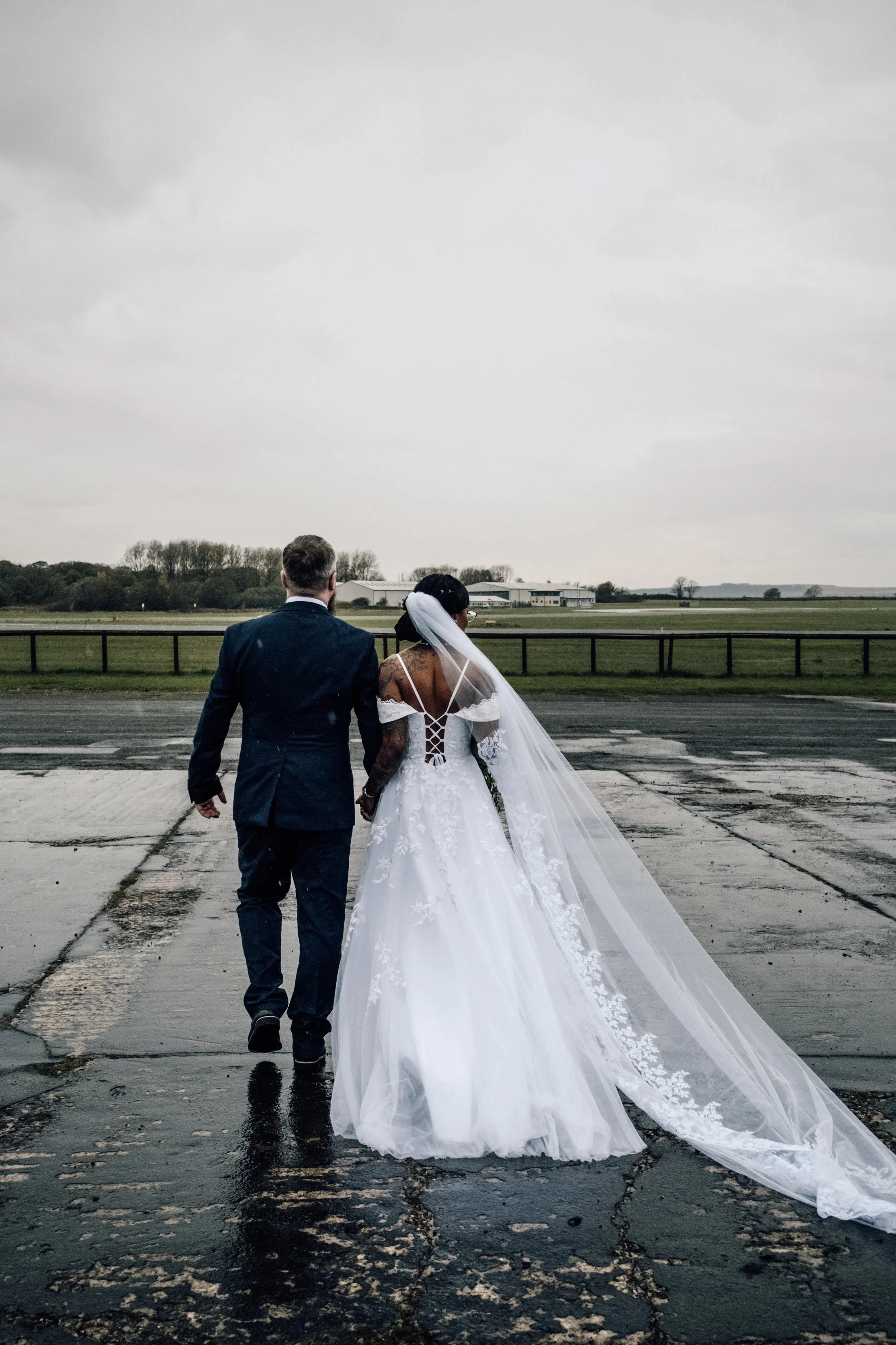 A bride and groom walking away on a wet airport tarmac, holding hands, with overcast sky and open fields in the background.