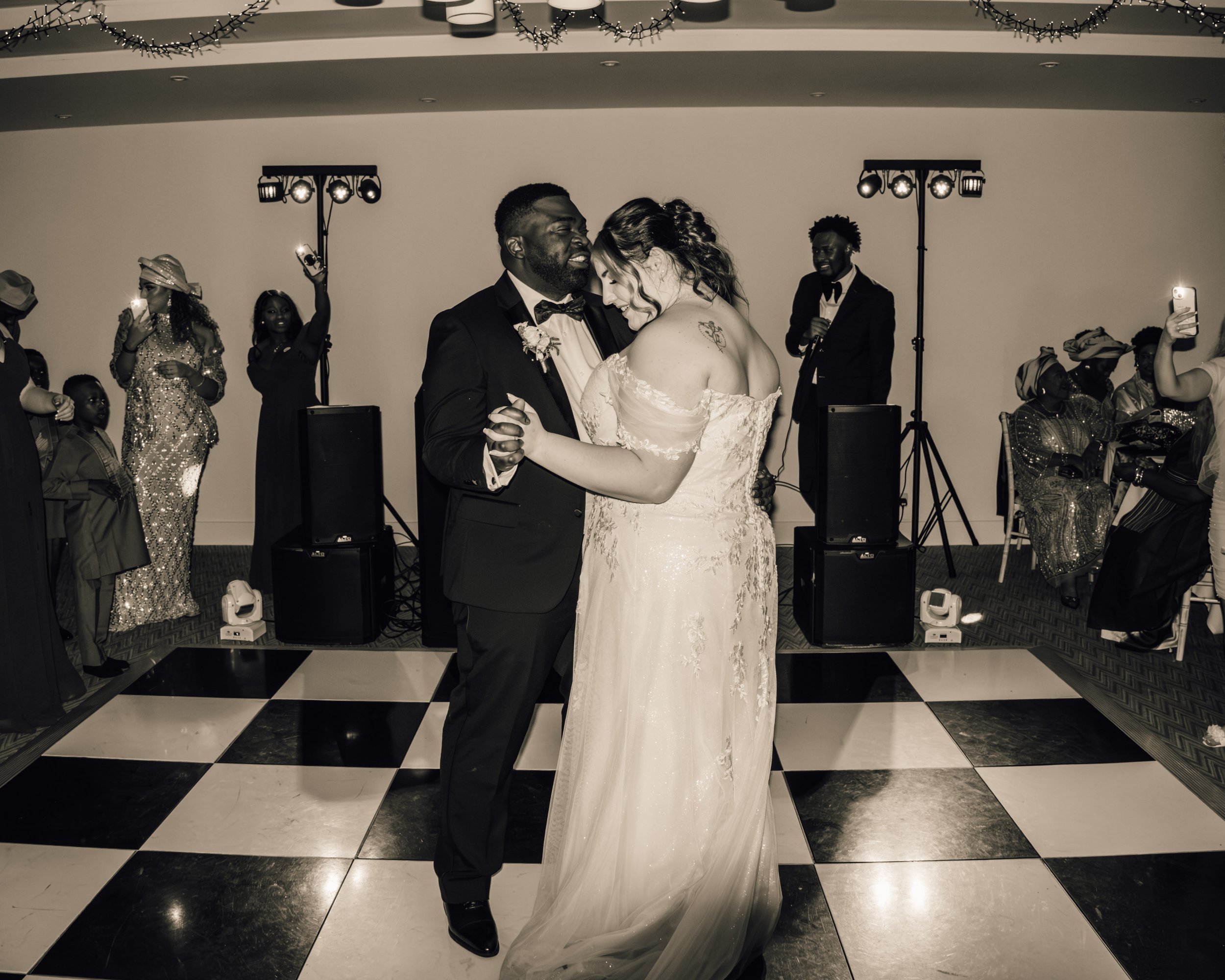 A newlywed couple dancing at their wedding reception, surrounded by guests, with a black-and-white checkered floor and decorative lighting.